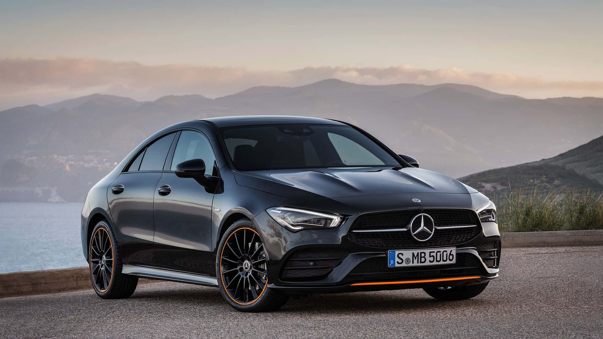 Mercedes Benz Cla-class 桌布 壁紙 — 一輛時尚的黑色平治CLA房車，帶有橙色點綴，停泊在日落時分俯瞰山巒和海洋的海岸公路上。