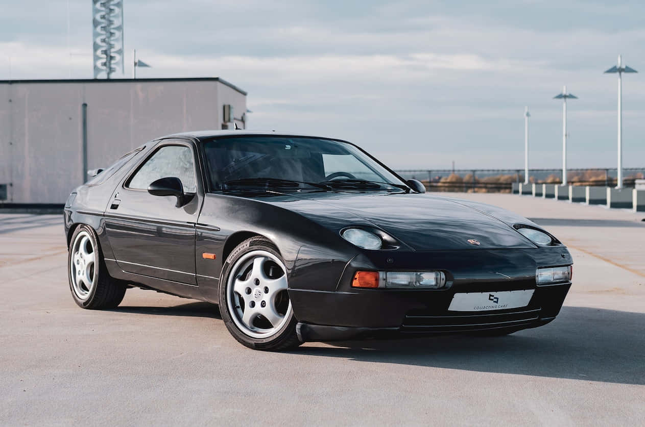Porsche 928 桌布 壁紙 — 一輛時尚的黑色保時捷928跑車停泊在混凝土停車場，天空多雲。