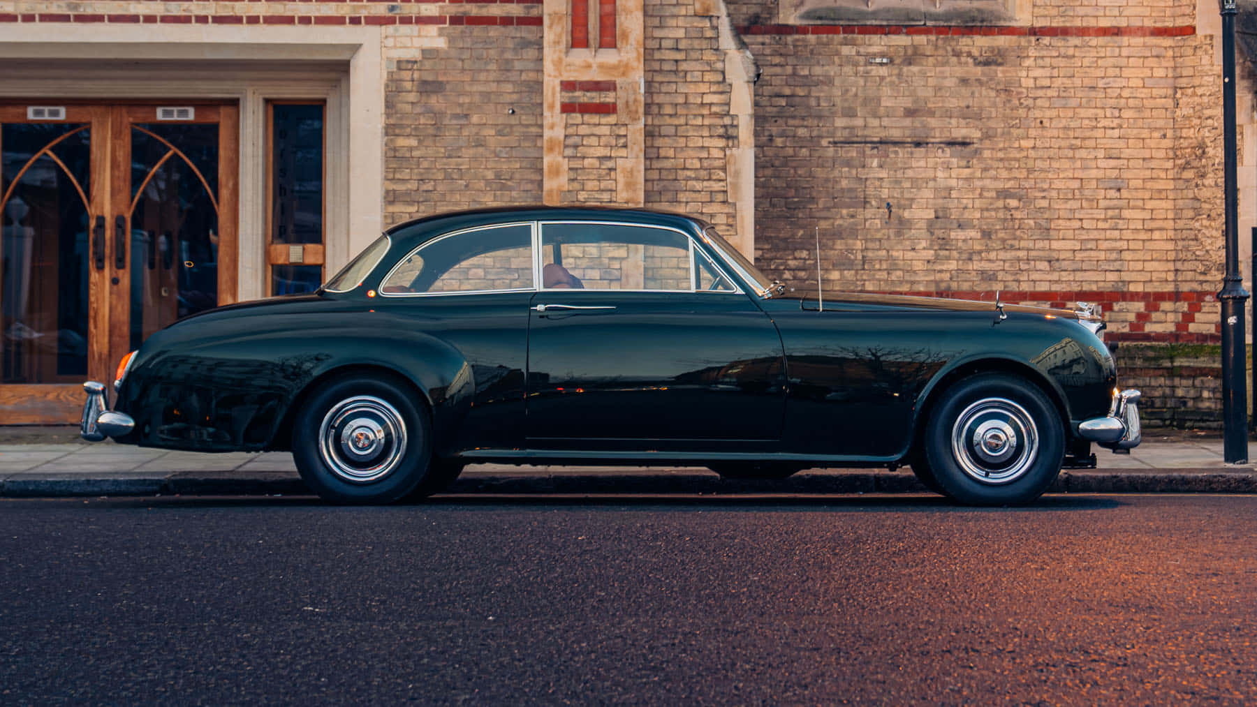 Bentley S2 桌布 壁紙 — 一輛時尚的黑色復古車停在陽光明媚的街道上，旁邊是帶有拱形木門的磚砌建築。