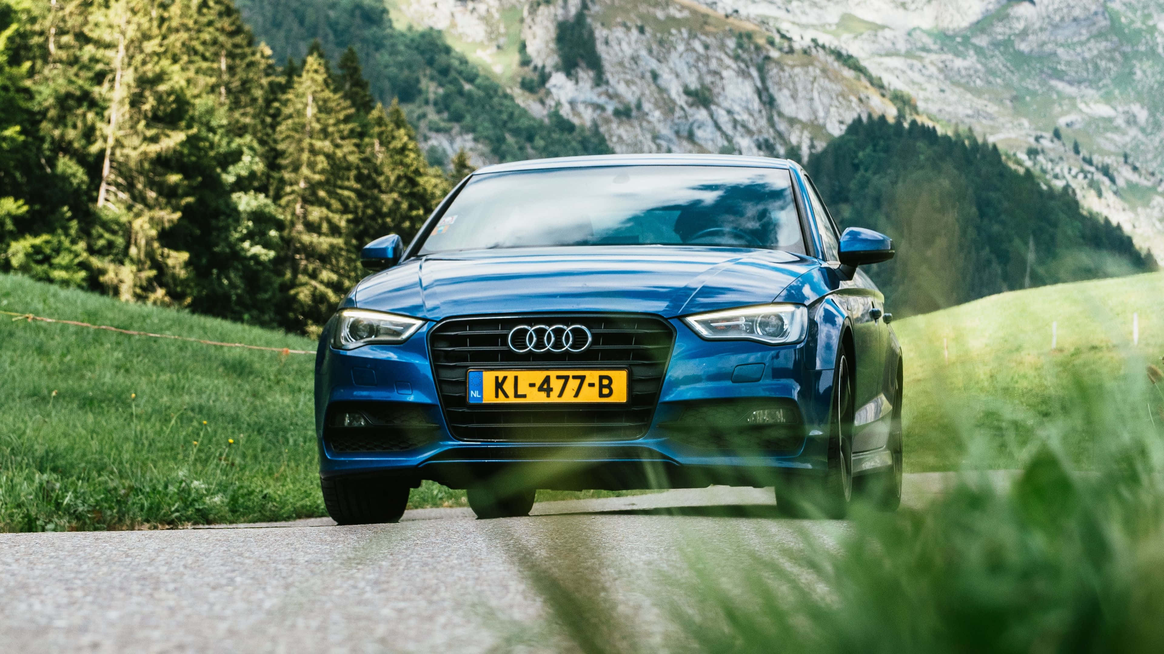 Audi Q7 桌布 壁紙 — 一架時尚的藍色奧迪房車沿著蜿蜒的山路行駛，周圍是茂密的綠色森林和岩石山峰。