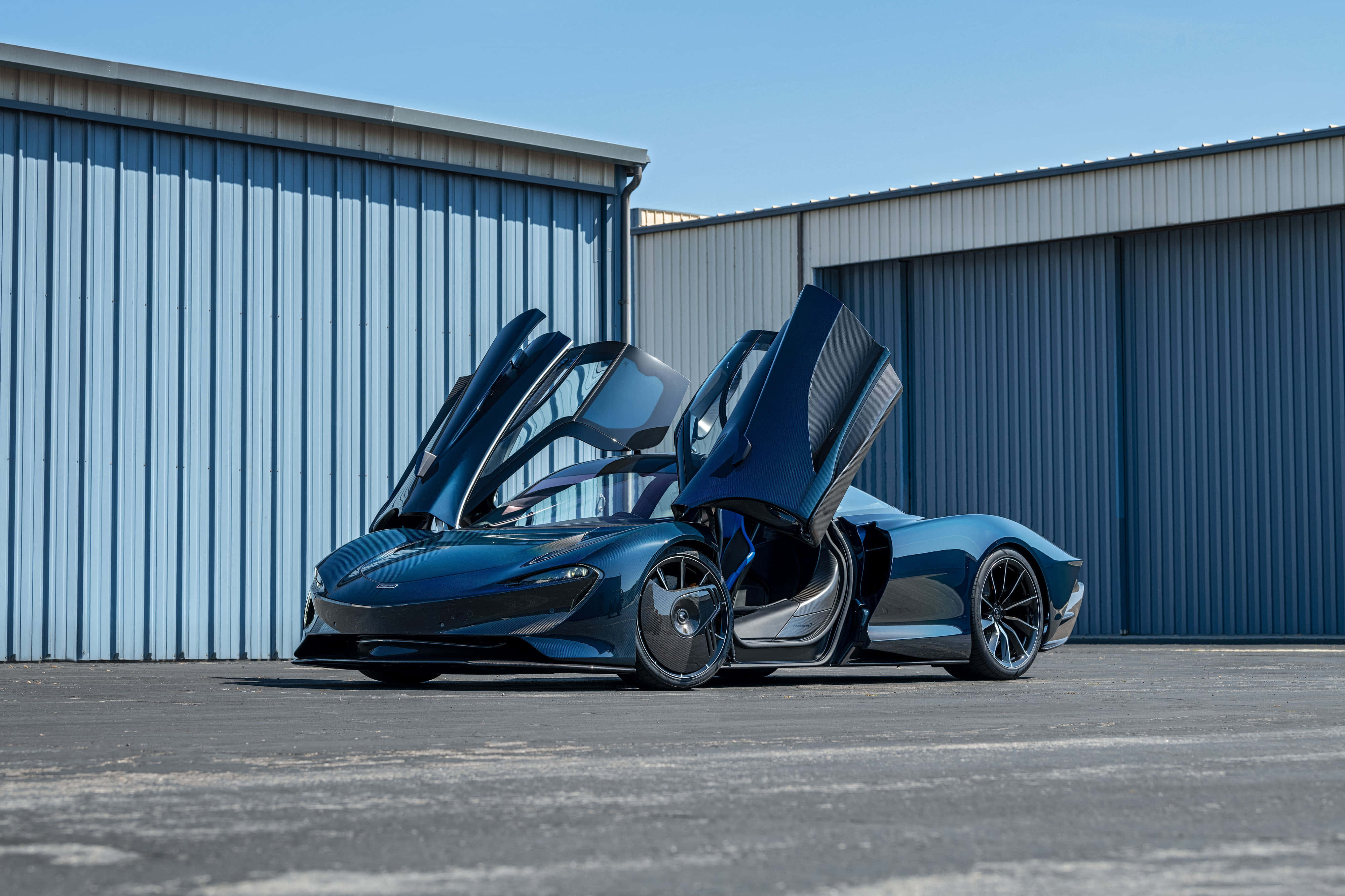 Mclaren Speedtail 桌布 壁紙 — 一架時尚的藍色跑車，鷗翼車門敞開，在晴朗的藍天下自豪地矗立在工業金屬倉庫前。