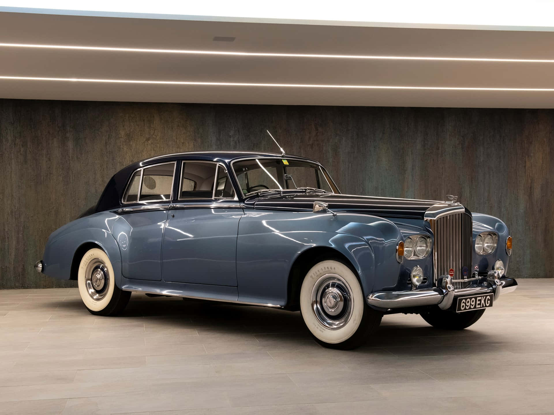 Bentley S3 桌布 壁紙 — 一輛時尚的藍色復古賓利轎車配備白色牆壁輪胎，在現代畫廊燈光下，以紋理木牆為背景閃閃發光。