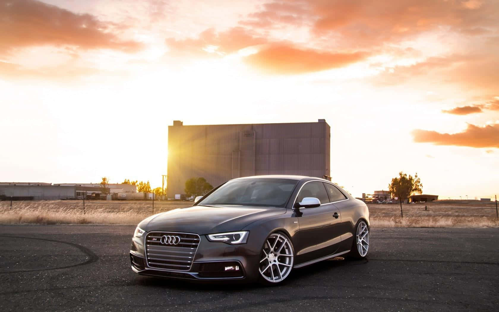 Audi S5 桌布 壁紙 — 一輛時尚的深灰色奧迪轎跑車在充滿活力的橙色日落天空下閃閃發光，背景是一座大型工業建築。