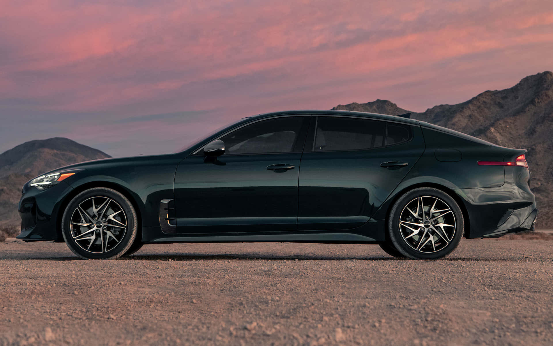 Kia Stinger 桌布 壁紙 — 一架時尚的深綠色轎車停泊在塵土飛揚的道路上，背景是山脈，天空是充滿活力的粉紅色和橙色日落。