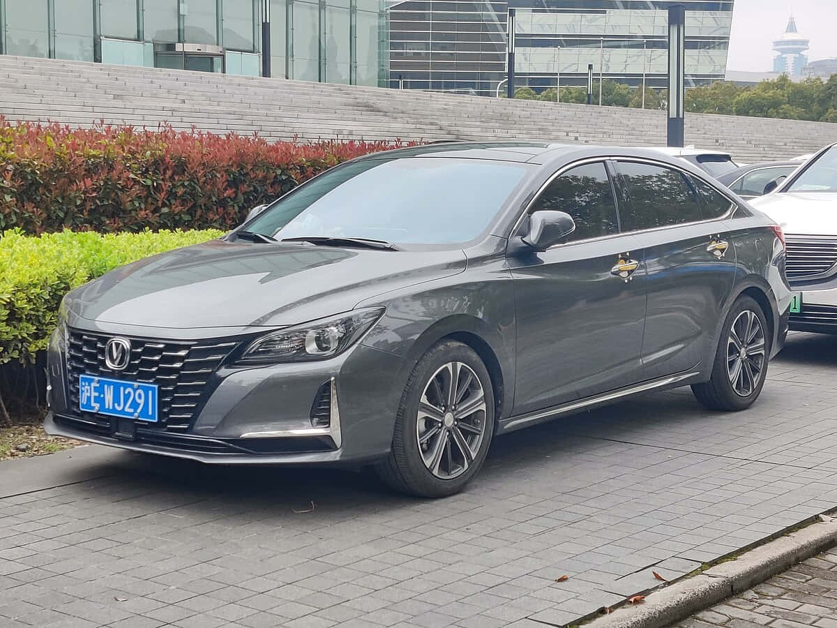  壁紙 — 一架時尚灰色房車停泊在鋪砌的城市街道上，旁邊有綠色灌木和現代玻璃建築。
