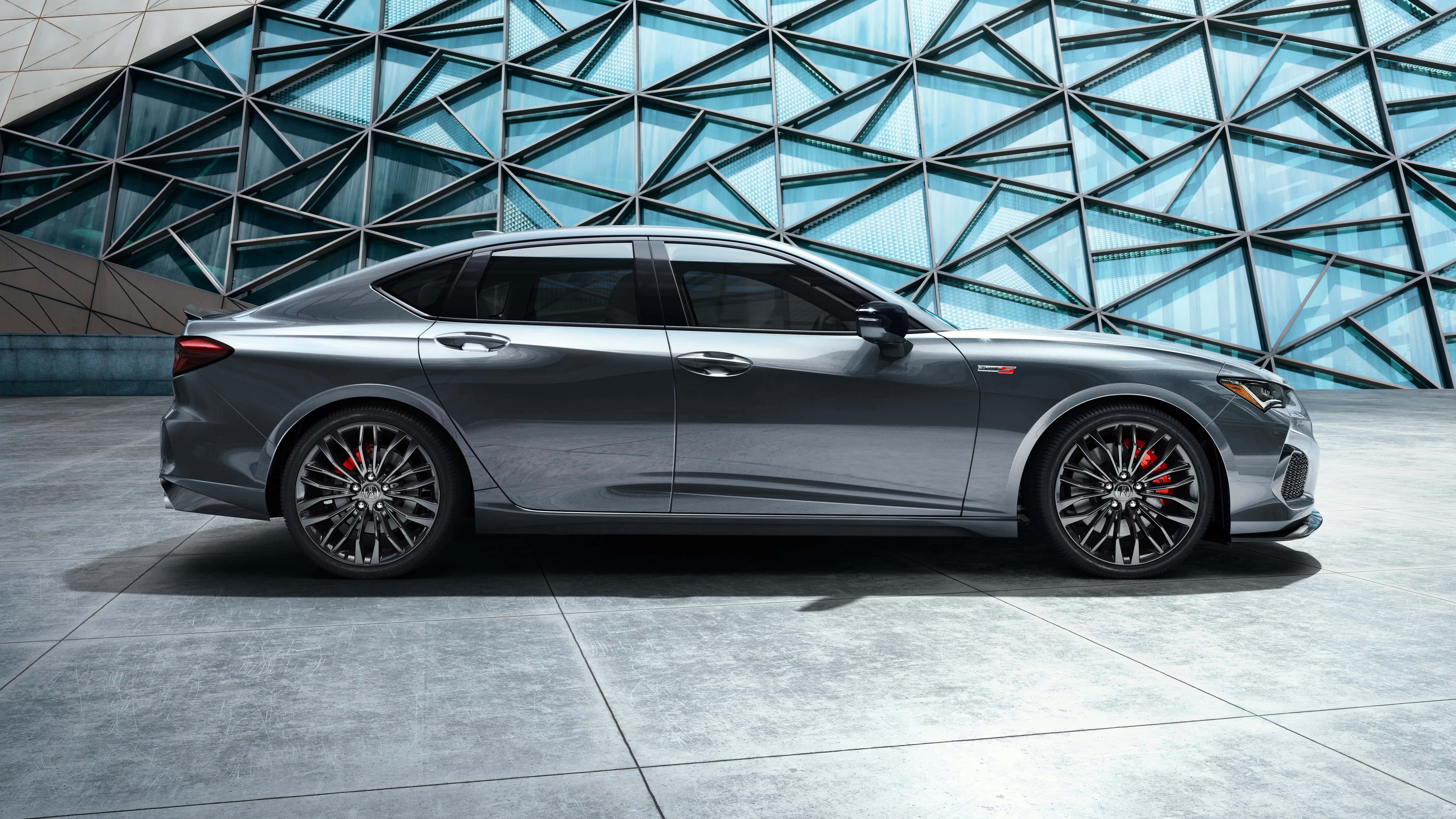 Acura Tlx 桌布 壁紙 — 一輛時尚的灰色運動轎車配備紅色制動卡鉗，停在一座未來主義幾何玻璃建築前。