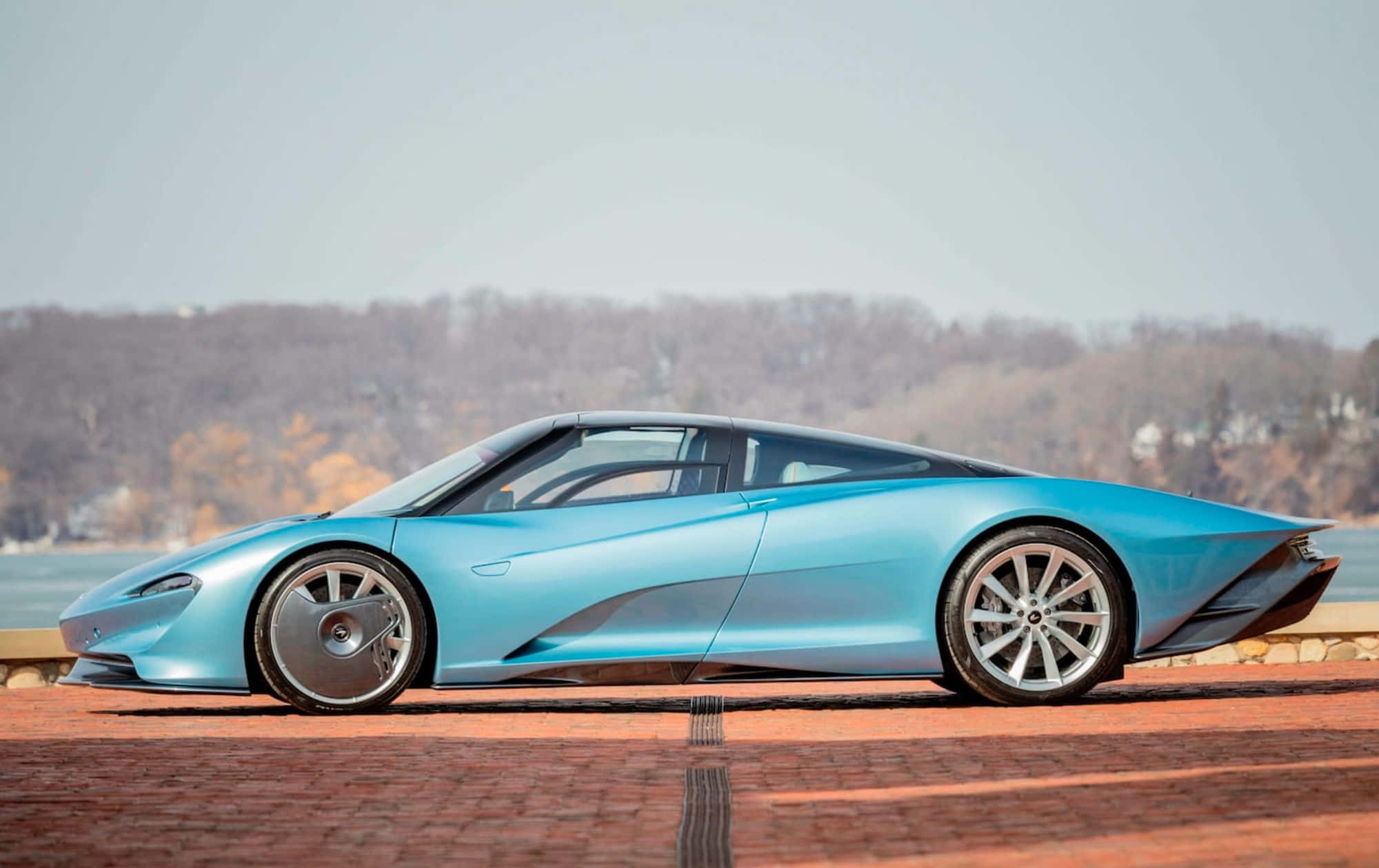 Mclaren Speedtail 桌布 壁紙 — 一架時尚淺藍色跑車停泊在磚路上，遠處有樹木成蔭的岸邊，天空朦朧。