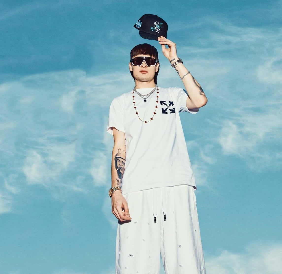  壁紙 — 一位穿著白色服裝和太陽眼鏡的時尚人士，在明亮的藍天和蓬鬆雲朵下傾斜黑色帽子。