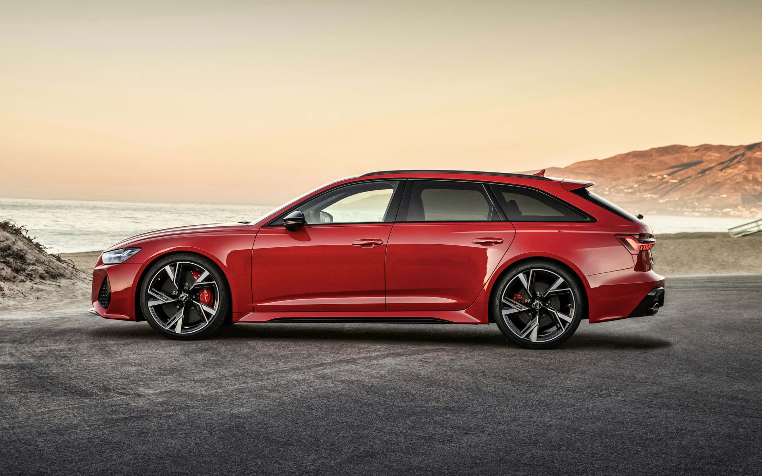 Audi Rs6 桌布 壁紙 — 一架時尚的紅色Audi RS6 Avant旅行車停泊在日落時分的海岸公路上，背景是海洋和山丘。