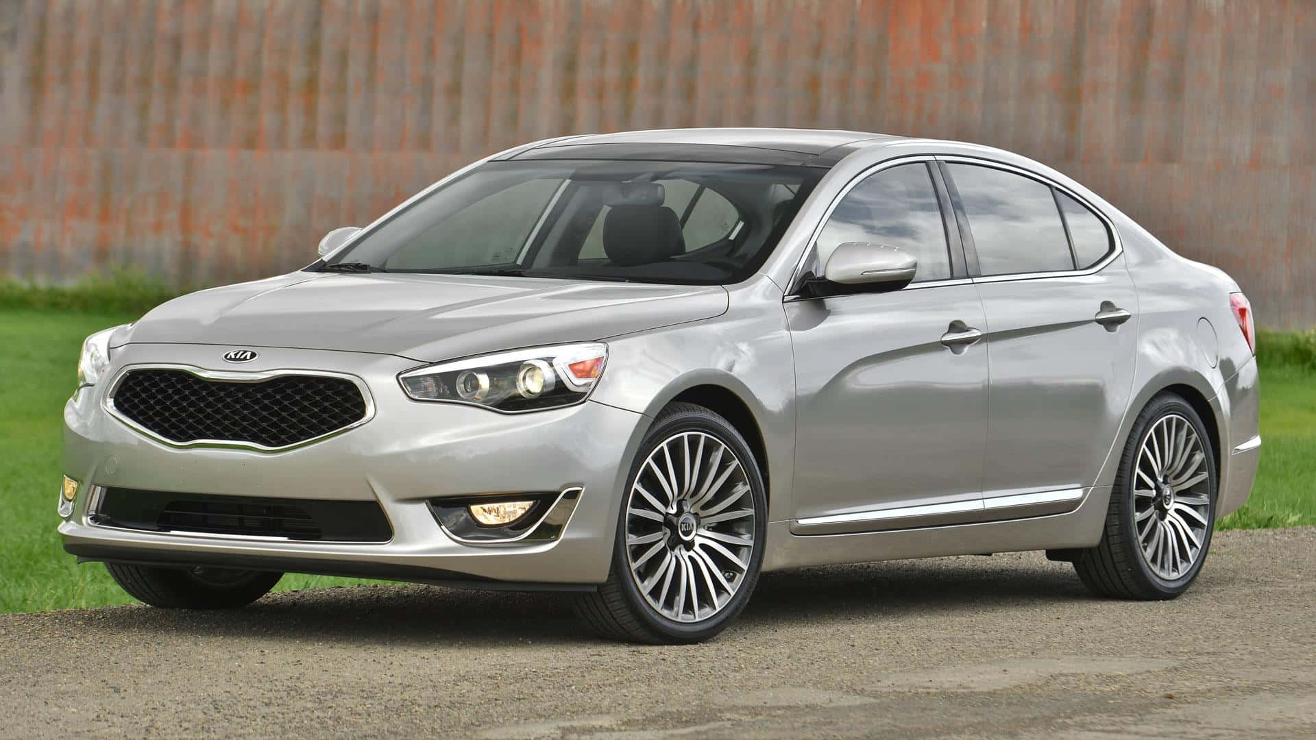 Kia Cadenza 桌布 壁紙 — 一輛時尚的銀色Kia轎車停在碎石路上，背景有草地和風化的木柵欄。