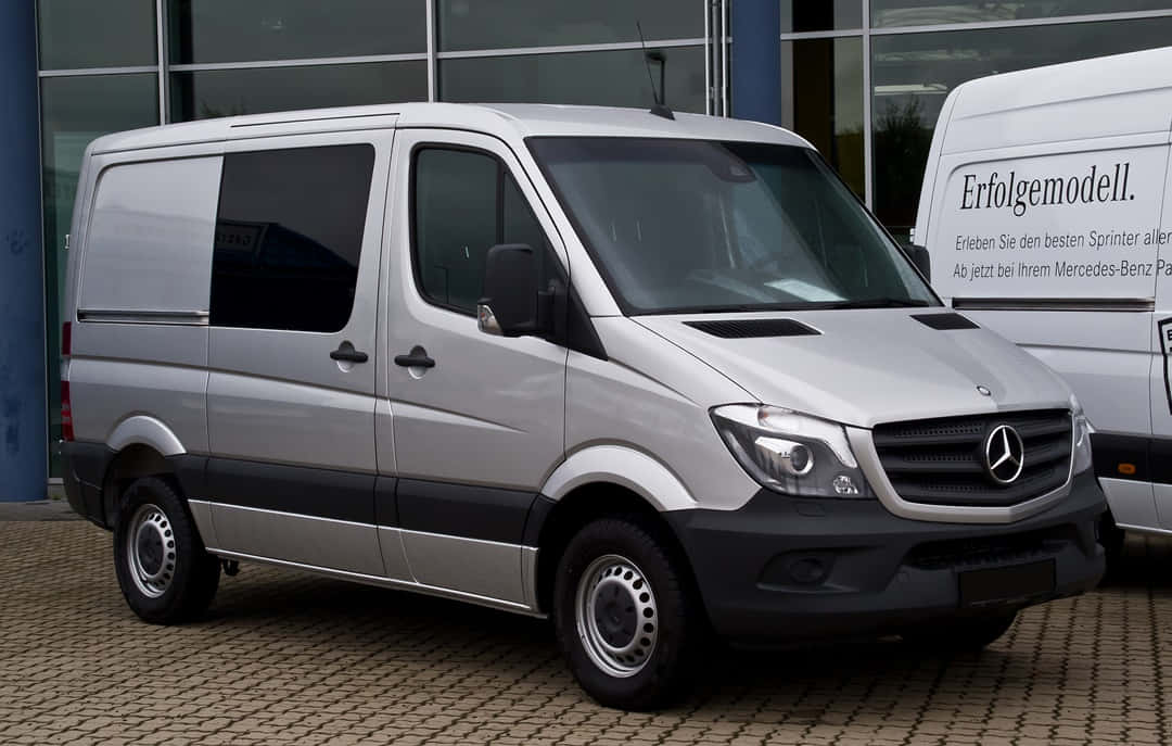 Mercedes Benz Sprinter 桌布 壁紙 — 一架時尚的銀色 Mercedes-Benz Sprinter 客貨車停泊在現代玻璃建築前的鋪砌停車場上。