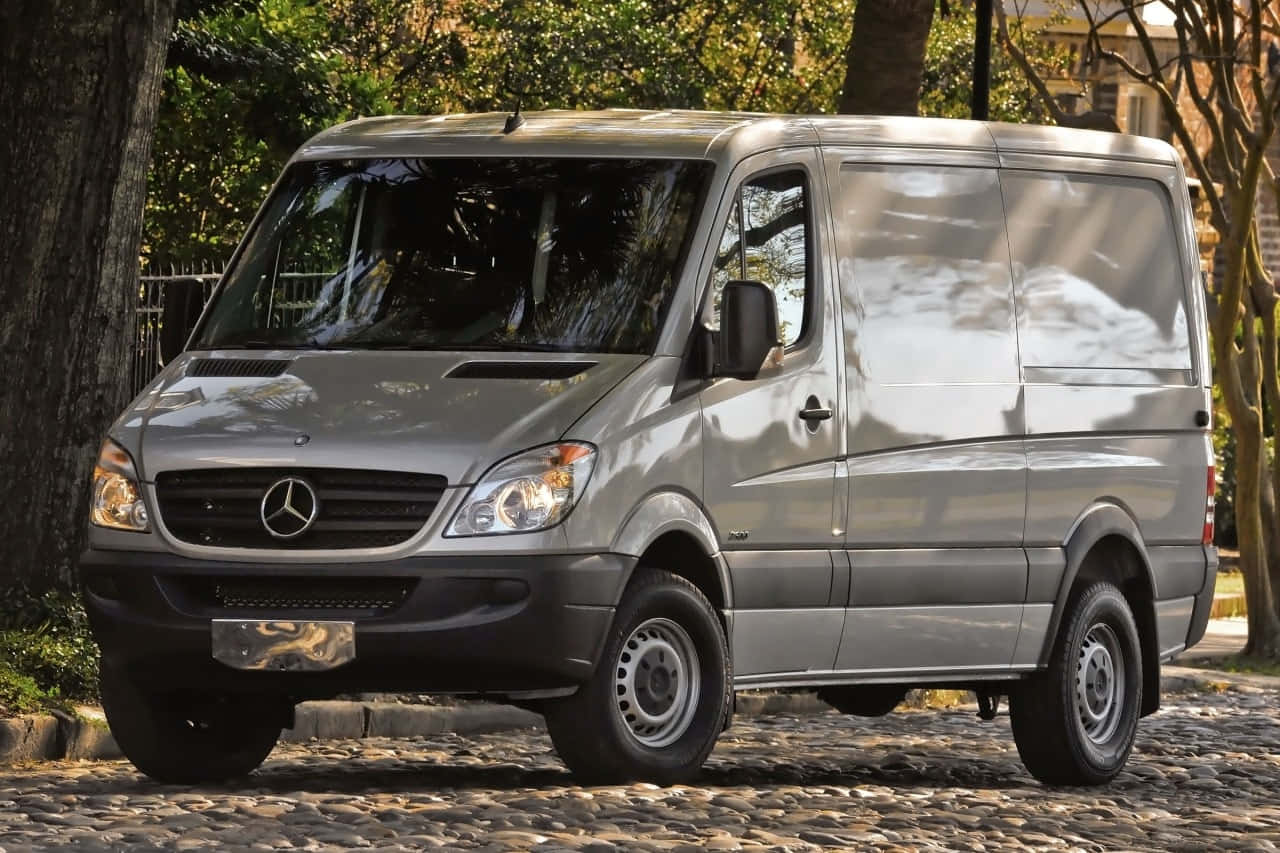 Mercedes Benz Sprinter 桌布 壁紙 — 一輛時尚的銀色梅賽德斯-奔馳 Sprinter 客貨車停泊在陽光普照、樹木成蔭的鵝卵石街道上。