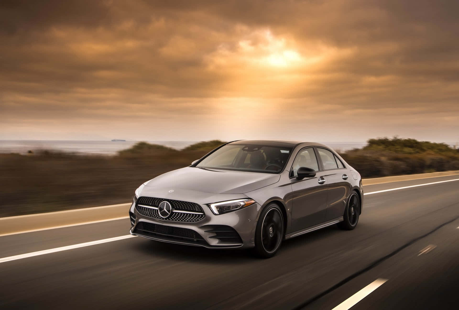 Mercedes Benz A-class 桌布 壁紙 — 一輛時尚的銀色梅賽德斯轎車在金色夕陽天空下的沿海高速公路上飛馳。