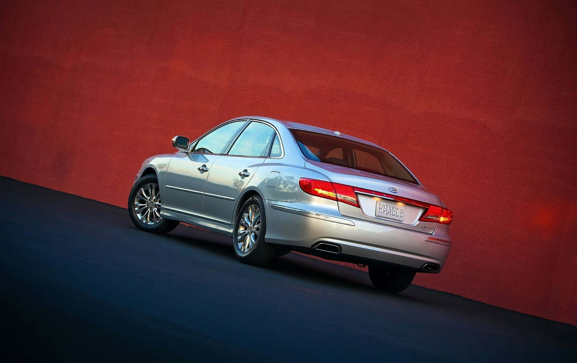 Hyundai Azera 桌布 壁紙 — 一輛時尚的銀色轎車在工作室燈光下傾斜靠在有紋理的紅色牆壁上。