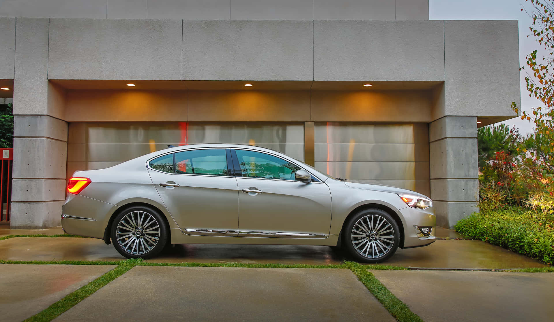Kia Cadenza 桌布 壁紙 — 一輛時尚的銀色轎車停在一棟現代混凝土建築前，建築內有溫暖的室內燈光。