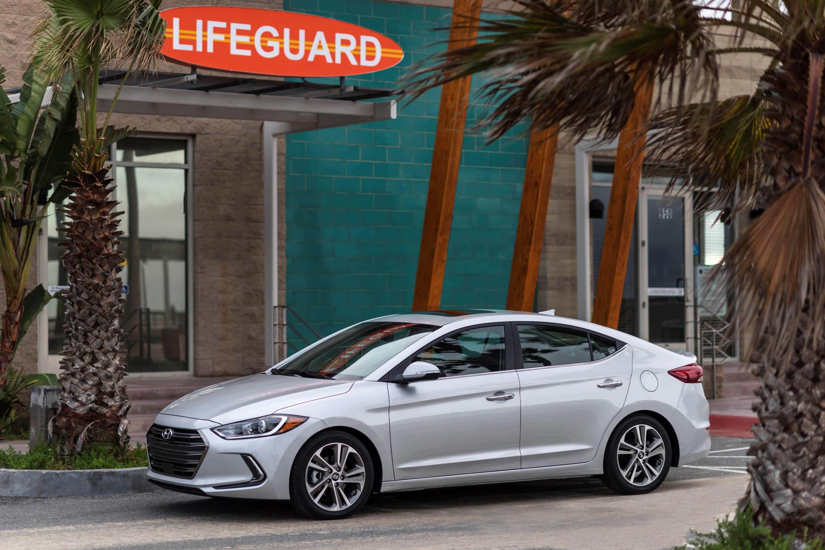 Hyundai Elantra 桌布 壁紙 — 一輛時尚的銀色轎車停泊在棕櫚樹環繞的藍綠色救生員站前。