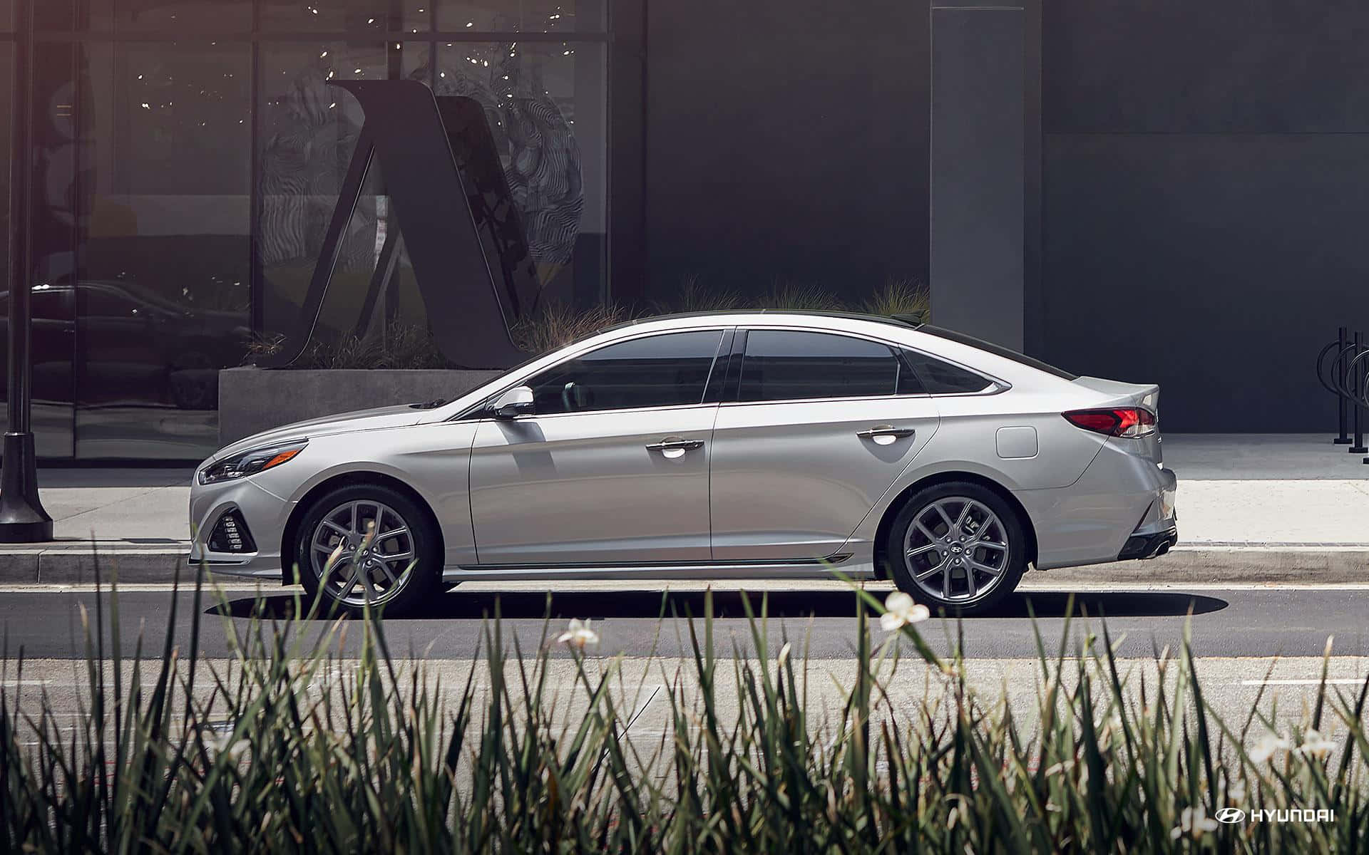 Hyundai Sonata 桌布 壁紙 — 一架時尚的銀色轎車停泊在現代玻璃建築和綠化前景的城市街道上。