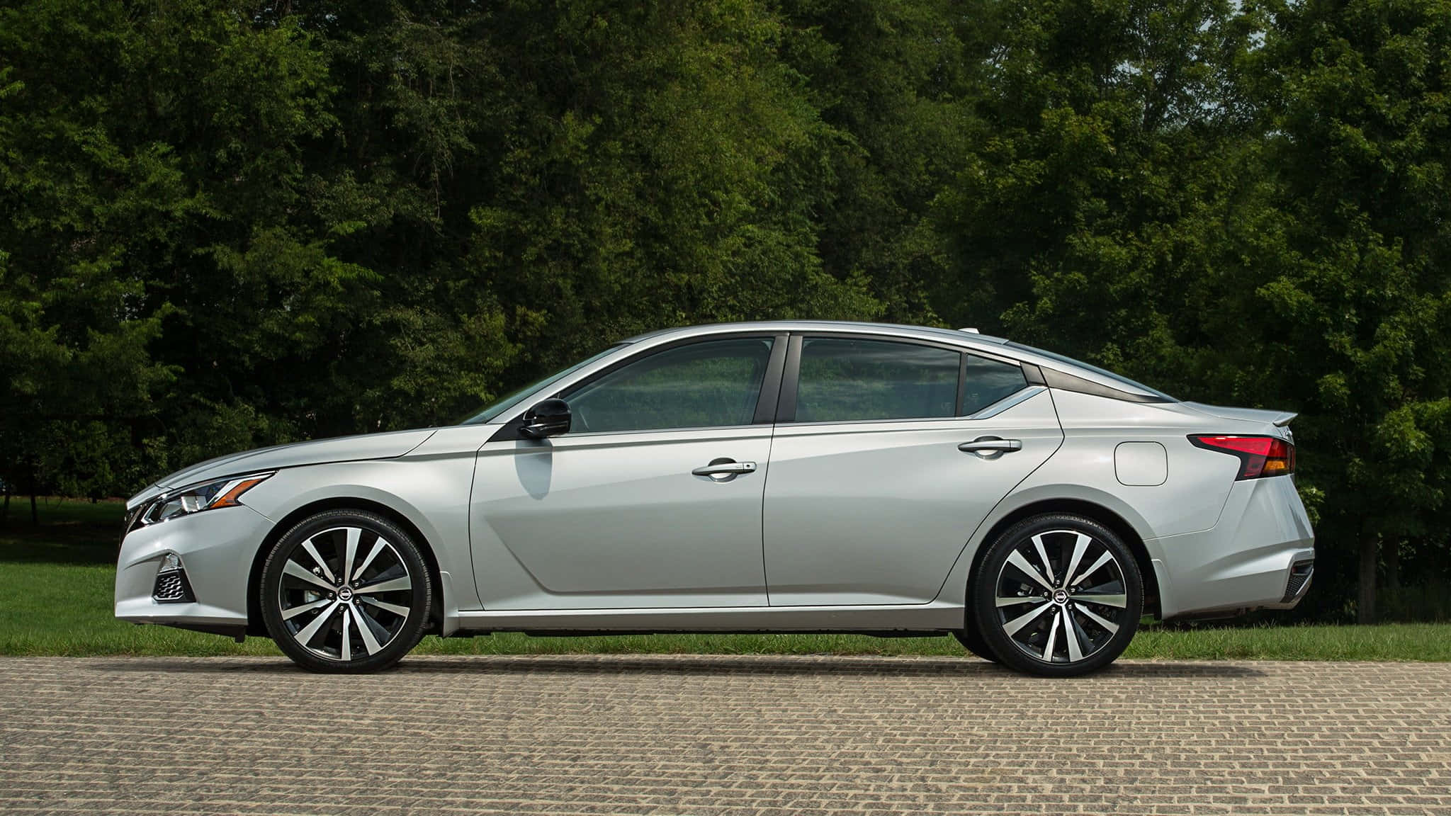 Nissan Altima 桌布 壁紙 — 一架時尚的銀色轎車停泊在鵝卵石小徑上，背景是茂密的綠樹。