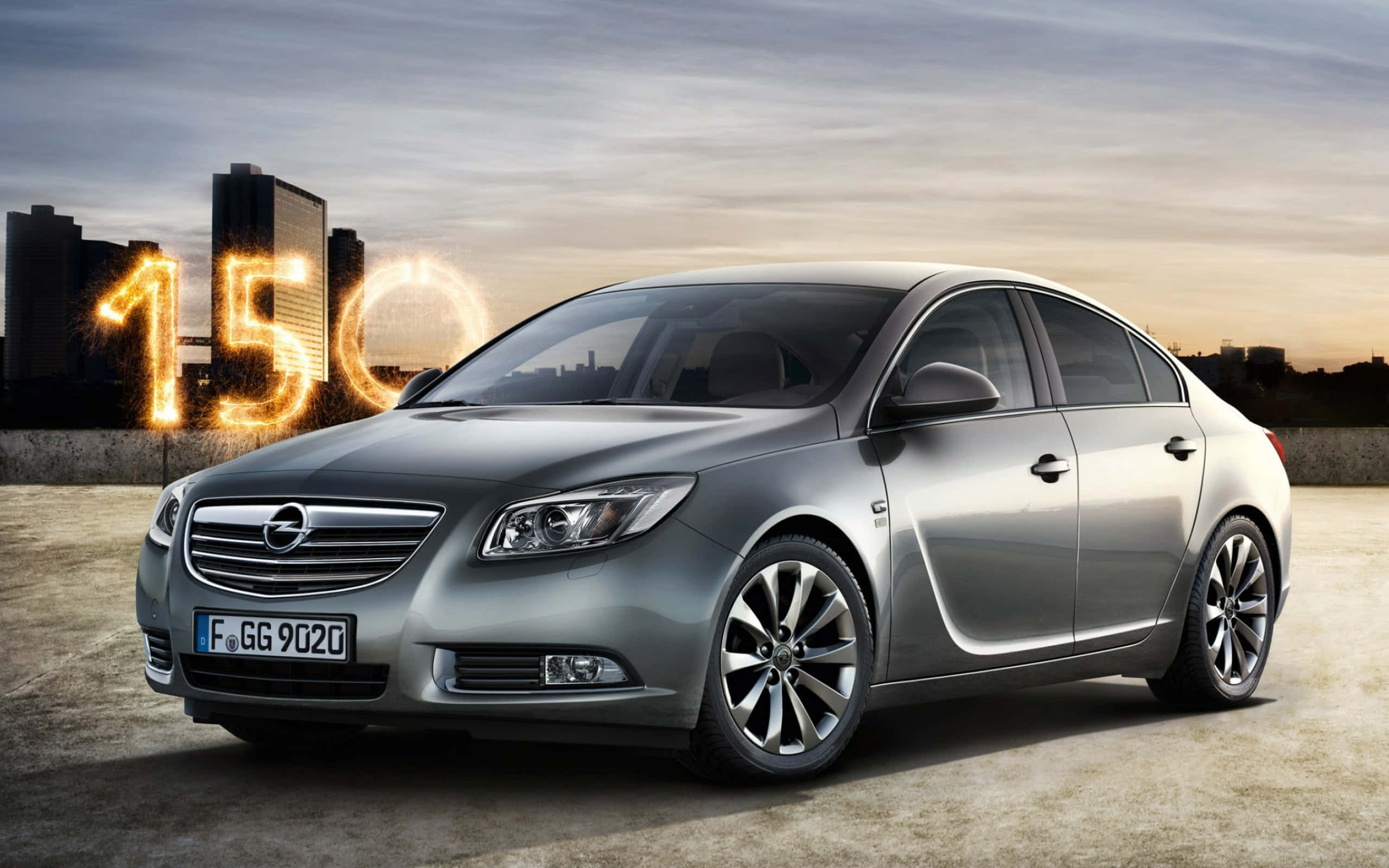 Opel Insignia 桌布 壁紙 — 一輛時尚的銀色轎車停在日落時分的屋頂上，背後是發光的「150」煙花，襯托著城市天際線。
