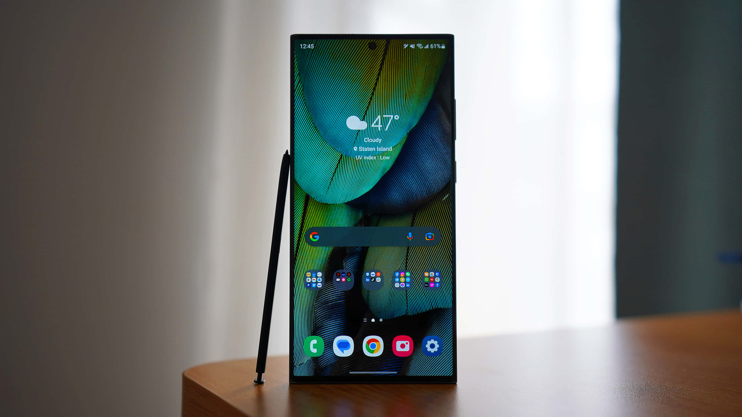 Samsung S23 Ultra 桌布 壁紙 — 一部時尚智能手機帶有鮮豔羽毛壁紙，直立於木桌上，旁邊放著觸控筆。