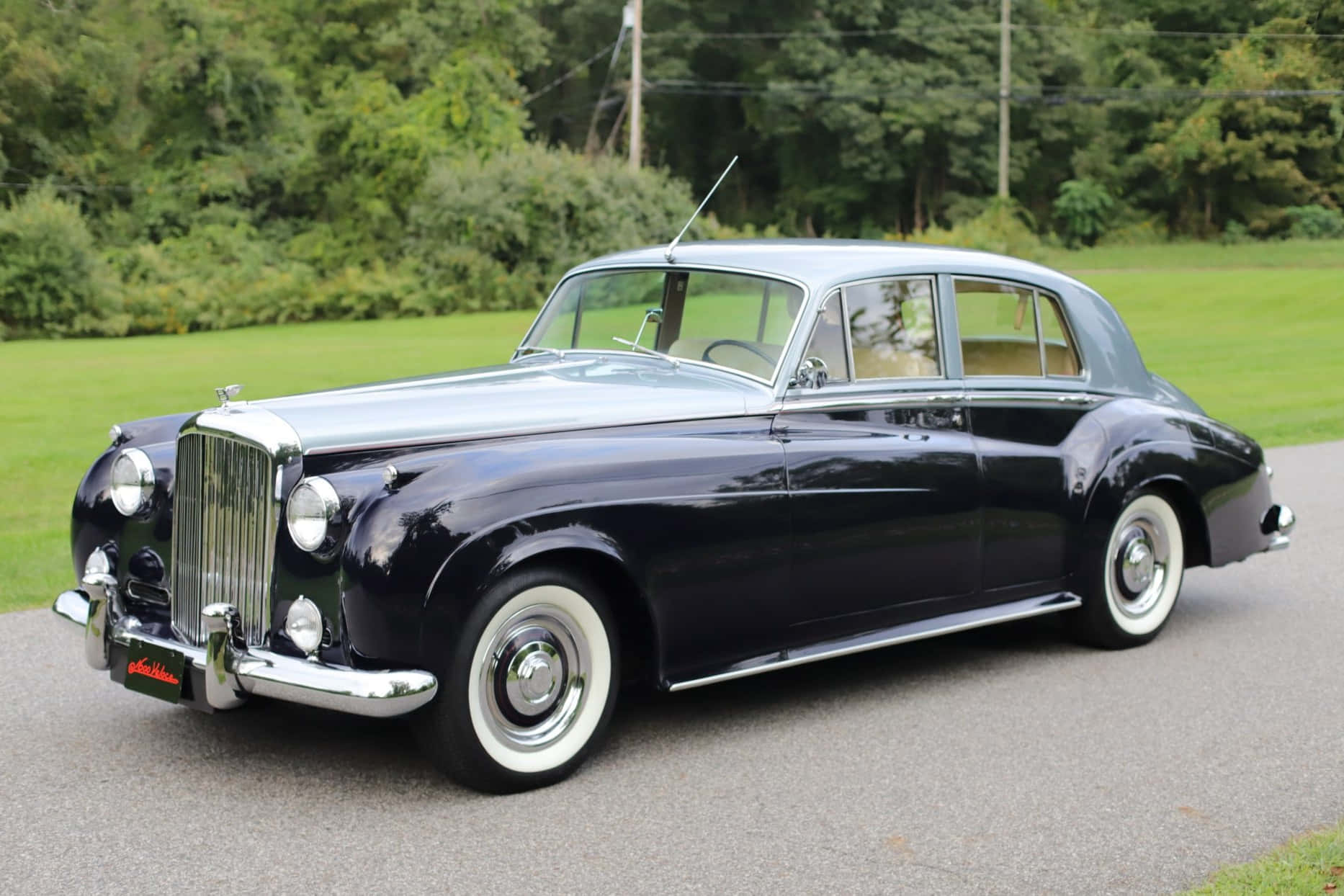 Bentley S2 桌布 壁紙 — 一輛時尚的復古賓利，採用黑色和銀色雙色調飾面，在日光下於一條兩旁長滿翠綠草地和樹木的瀝青路上閃閃發光。