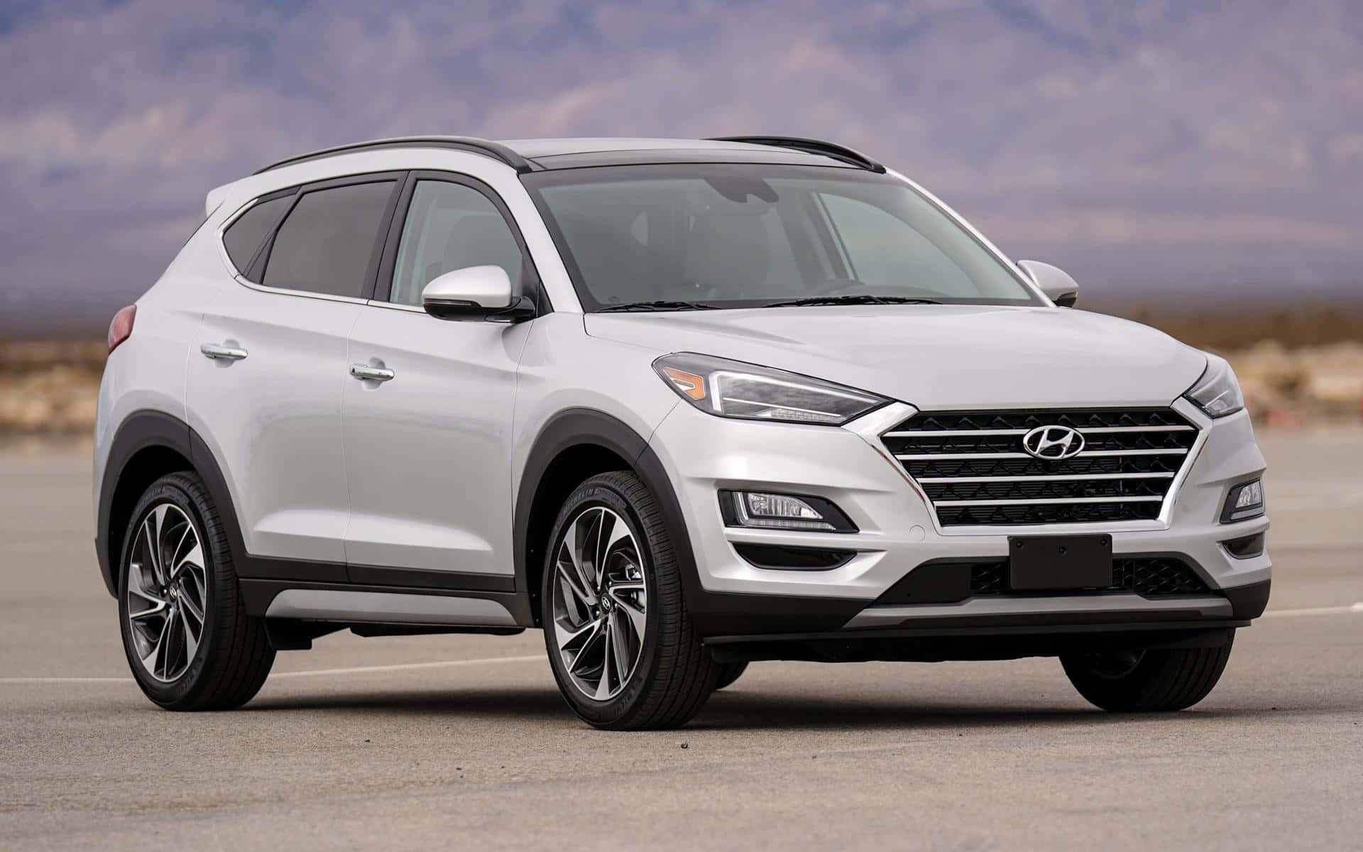 Hyundai Tucson 桌布 壁紙 — 一架時尚的白色現代 Tucson SUV 停泊在鋪砌的停車場上，背景是朦朧的山巒，天空多雲。