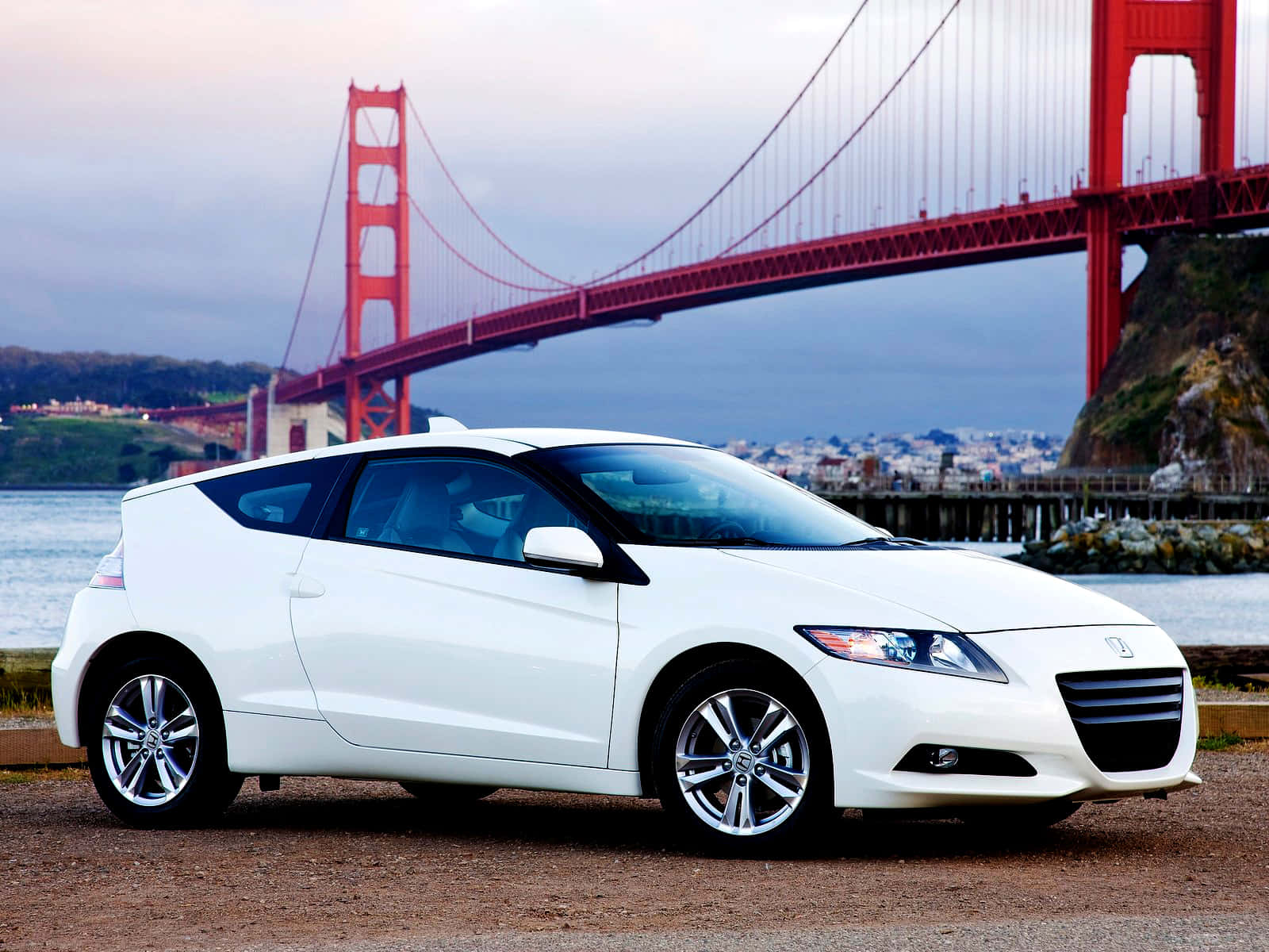 Honda Cr-z 桌布 壁紙 — 一架時尚的白色跑車停在水邊，背景是標誌性的紅色金門大橋在陰雲密布的天空下聳立。
