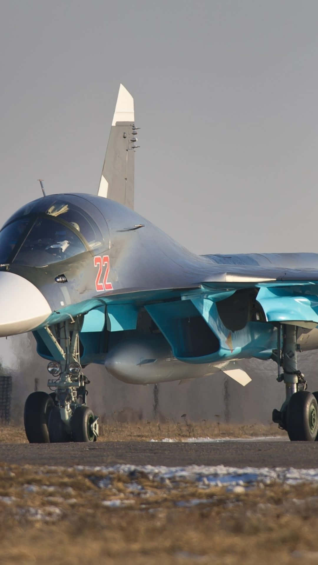 藍色蘇霍伊Su-34安卓巨無霸背景 壁紙 — 一架時尚的藍灰色戰鬥機，帶有紅色22號，停在蒼白天空下的雪地跑道上。