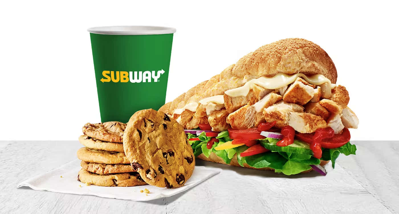 Subway 桌布 壁紙 — Subway 雞肉三文治配新鮮蔬菜，旁邊放著一個綠色飲料杯，還有一疊朱古力曲奇餅，放在白色紋理表面上。