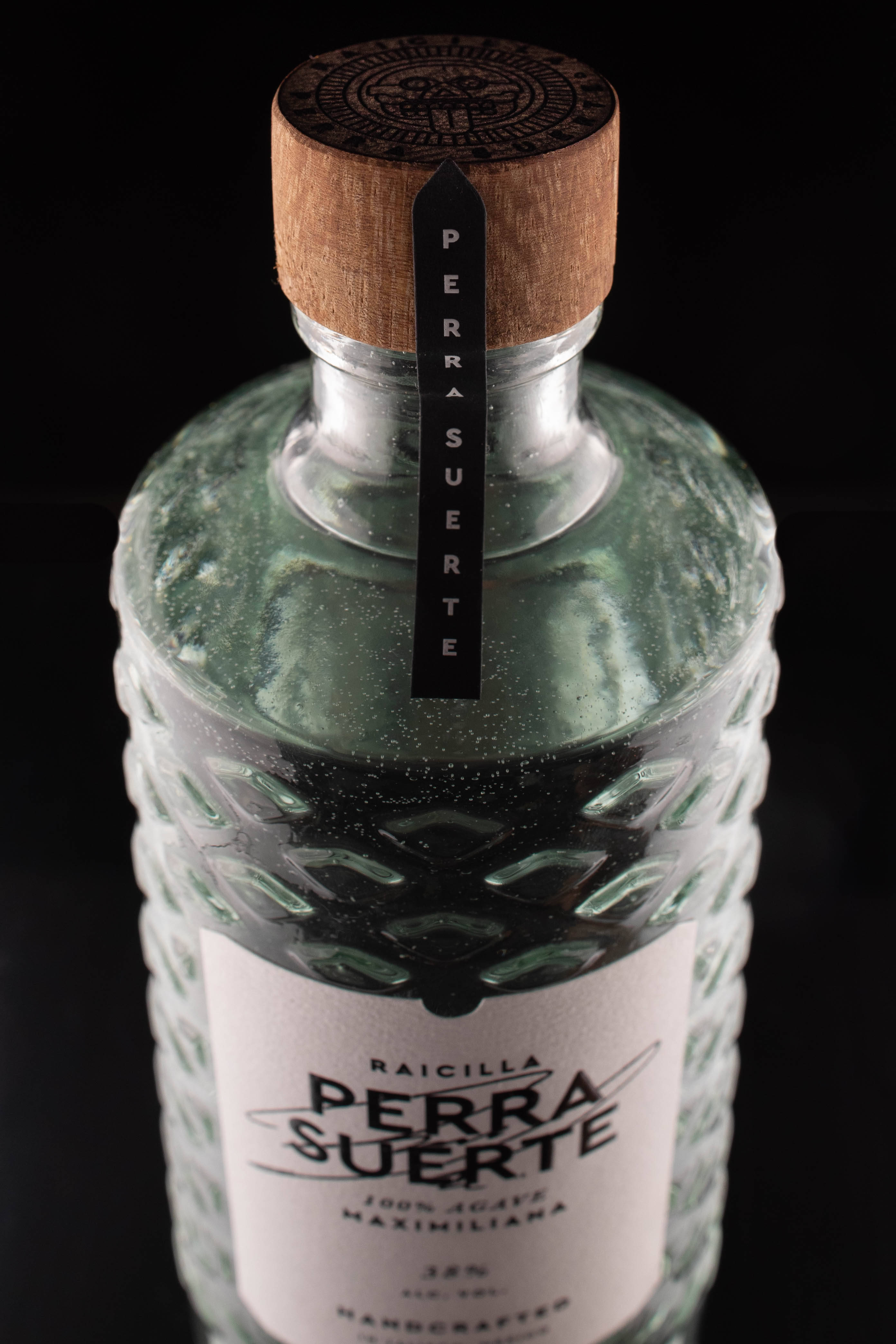Suerte Raicilla Perra龍舌蘭酒瓶 壁紙 — 特寫鏡頭：一個帶有木蓋的紋理綠色玻璃瓶Raicilla Perra Suerte，置於黑色背景前。