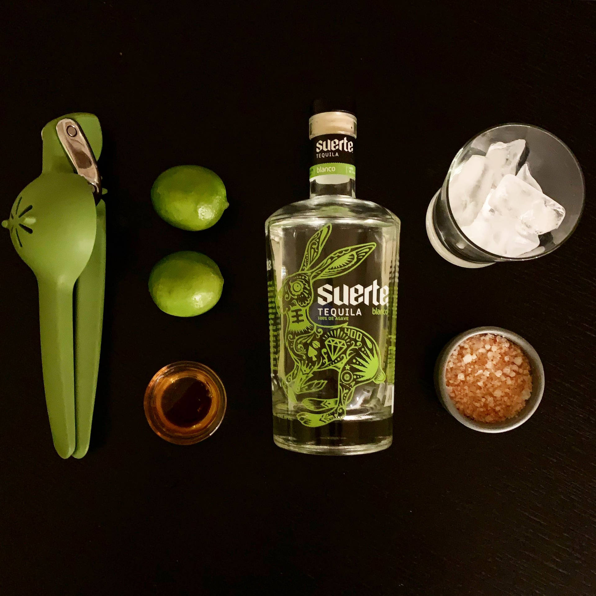 龍舌蘭酒瓶搭配青檸、青檸壓榨器、鹽和冰塊，置於深色木頭上。