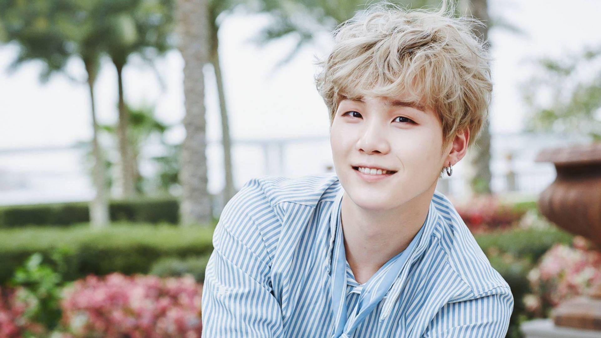 可愛休閒Suga BTS 壁紙 — 一位頭髮蓬鬆的金髮年輕男子在陽光普照的花園中，棕櫚樹和粉紅色花朵的襯托下，溫暖地微笑。