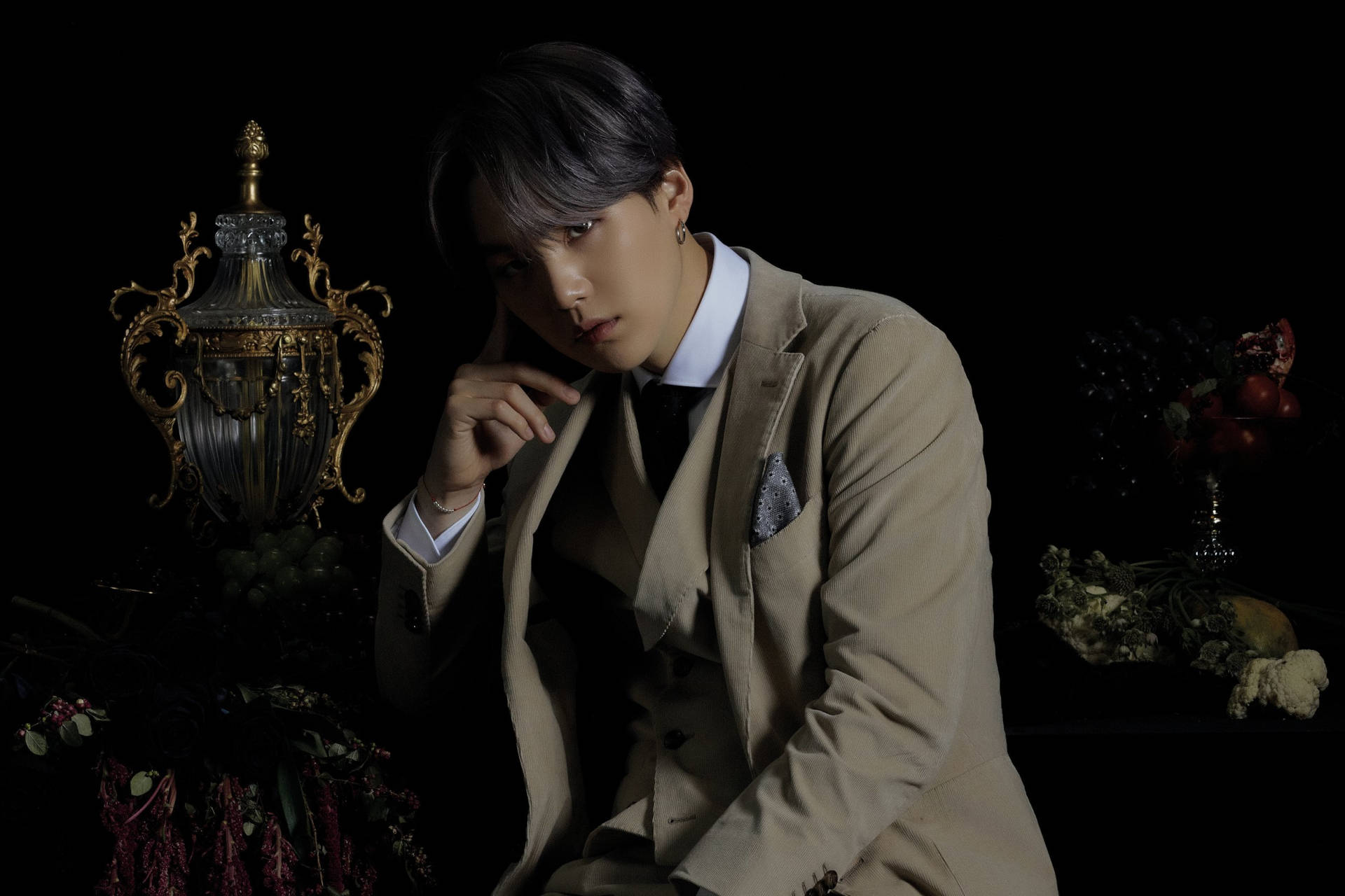 Suga BTS 深棕西裝造型 壁紙 — 一名身穿米色西裝的男子在深色背景下，帶有華麗金色裝飾和紅花，若有所思地擺姿勢。