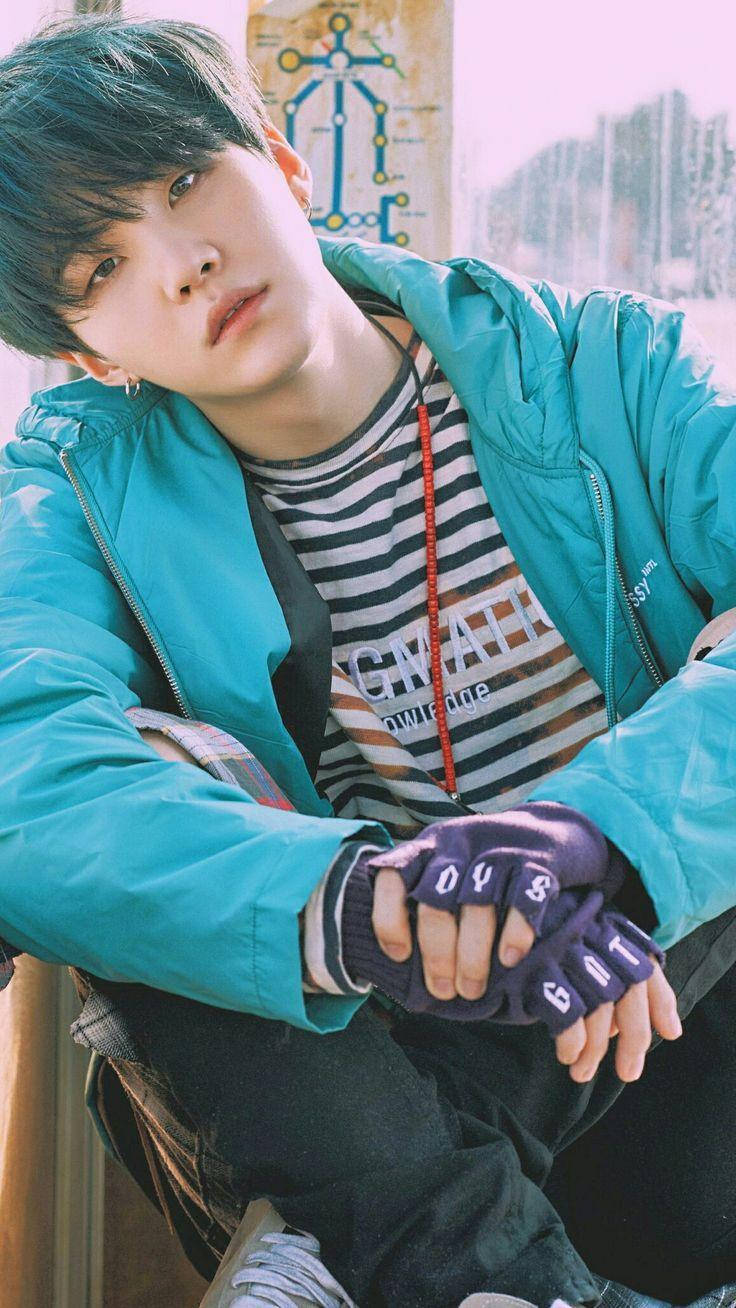 Suga BTS 湊綠外套 壁紙 — 一位青綠色頭髮的年輕男子靠著牆，身穿亮藍色外套和無指手套。