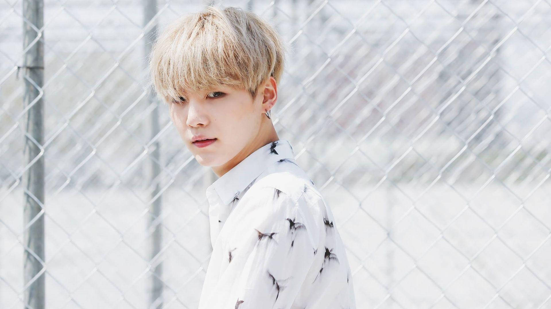 Suga BTS 白金髮型 壁紙 — 一位金髮青年穿著星星圖案襯衫，在明亮的日光下站在鐵絲網圍欄前。