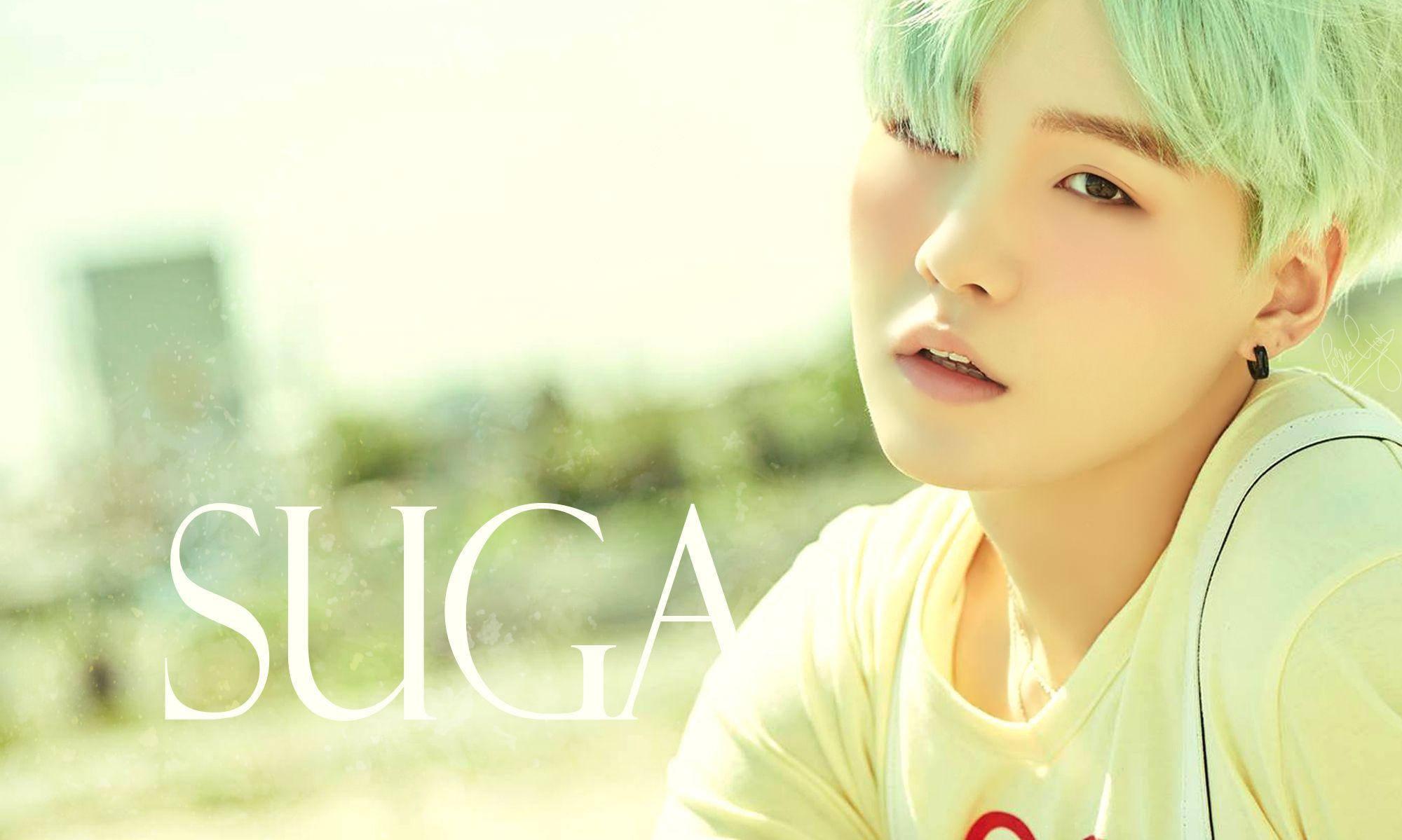 帥氣Suga BTS 壁紙 — 一位擁有薄荷綠髮色的年輕人，在明亮朦朧的背景上柔和地凝視，上面疊加了SUGA的名字。