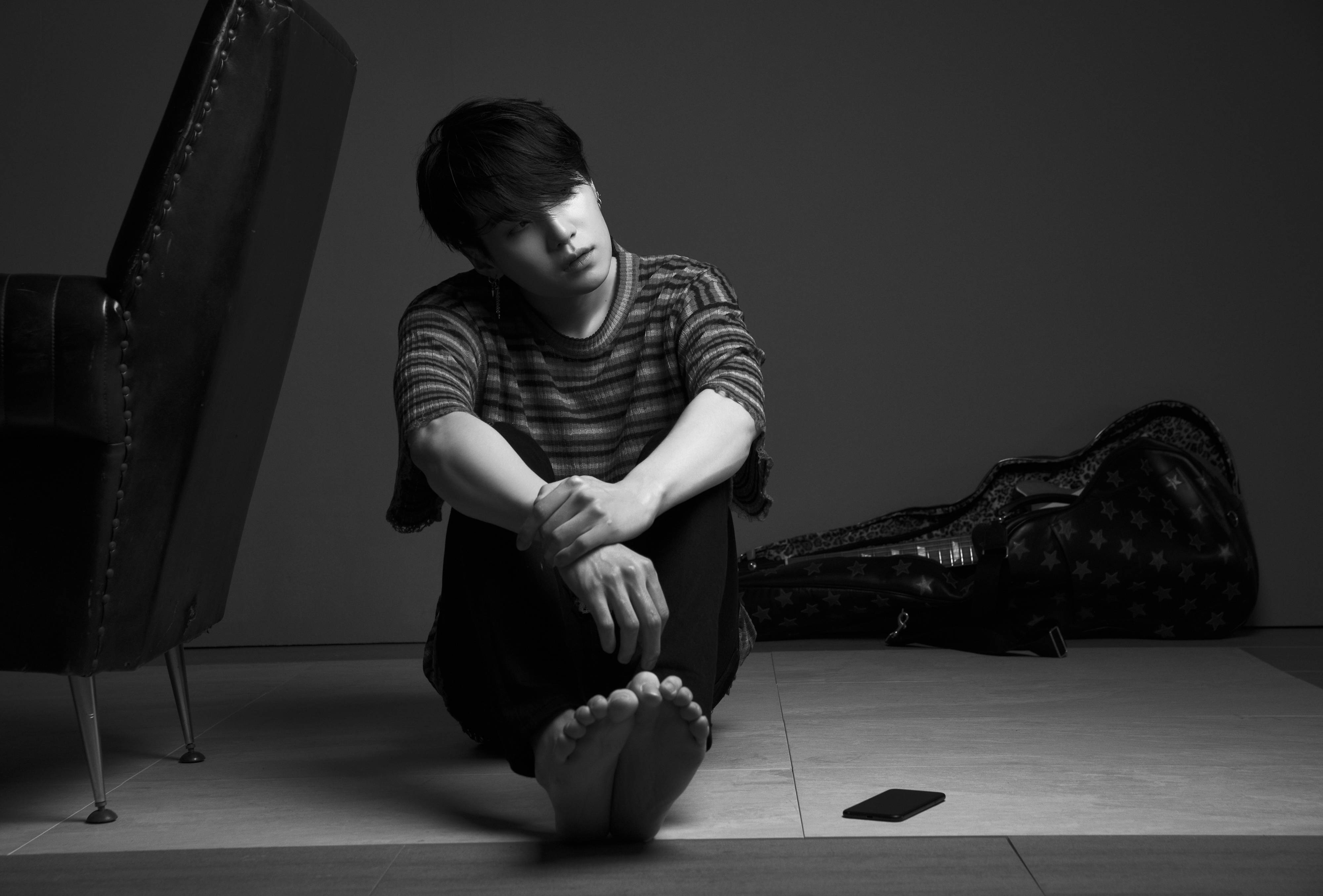 Suga BTS 單色系造型 壁紙 — 一名年輕男子赤腳坐在瓷磚地板上，旁邊放著結他盒和扶手椅，是一張清晰的黑白工作室照片。