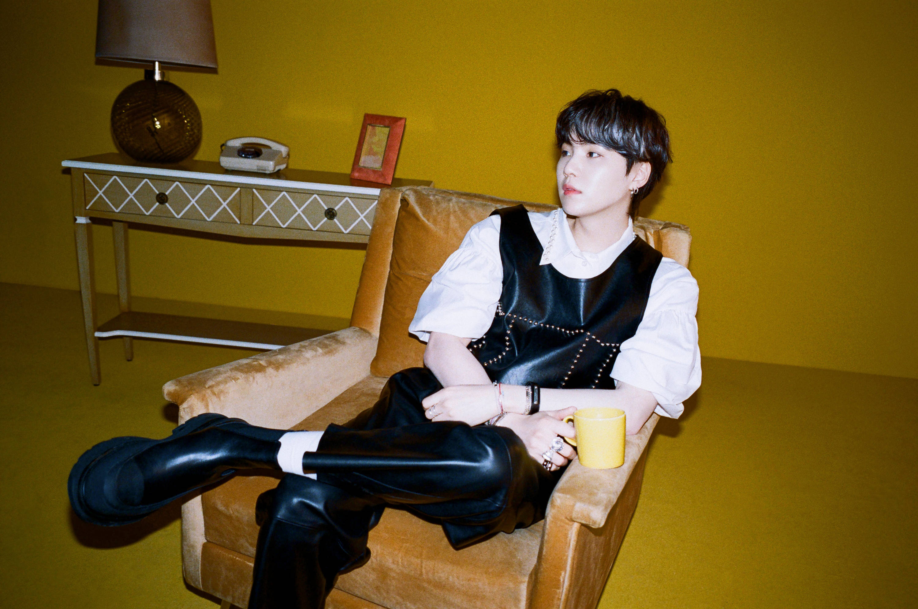 Suga BTS《Butter》 壁紙 — 一名年輕男子懶洋洋地坐在芥末黃色扶手椅上，手持黃色馬克杯，背景是充滿活力的黃色牆壁，帶有復古裝飾。