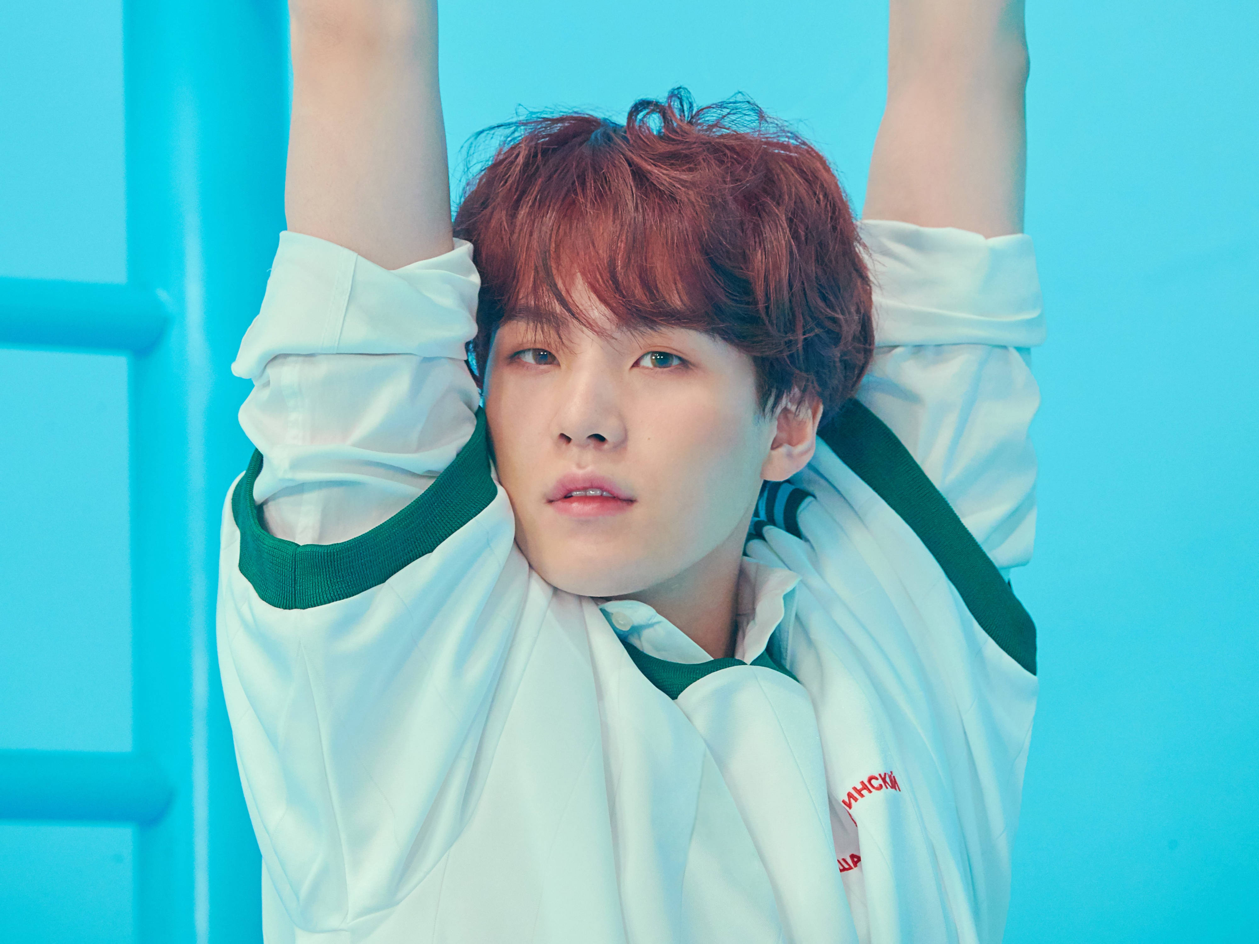 Suga BTS《Love Yourself: Answer》 壁紙 — 一位紅髮年輕男子穿著白色外套，在鮮豔的藍色背景前向上伸展雙臂。