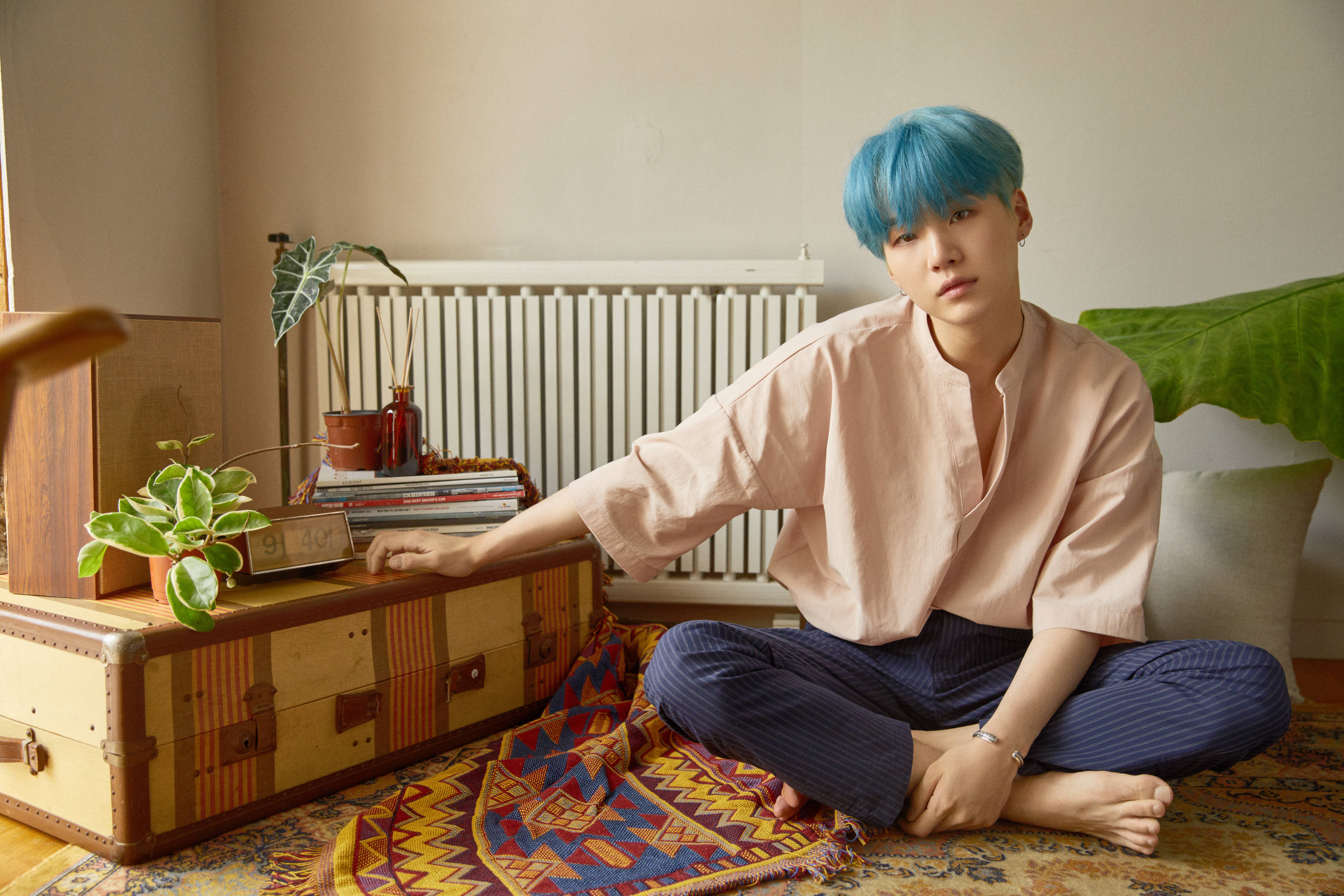 Suga BTS《Love Yourself: Her》概念照 壁紙 — 一個擁有充滿活力的藍色頭髮的人，在溫暖照明的房間裡，盤腿坐在有圖案的地毯上，旁邊放著一個復古手提箱。