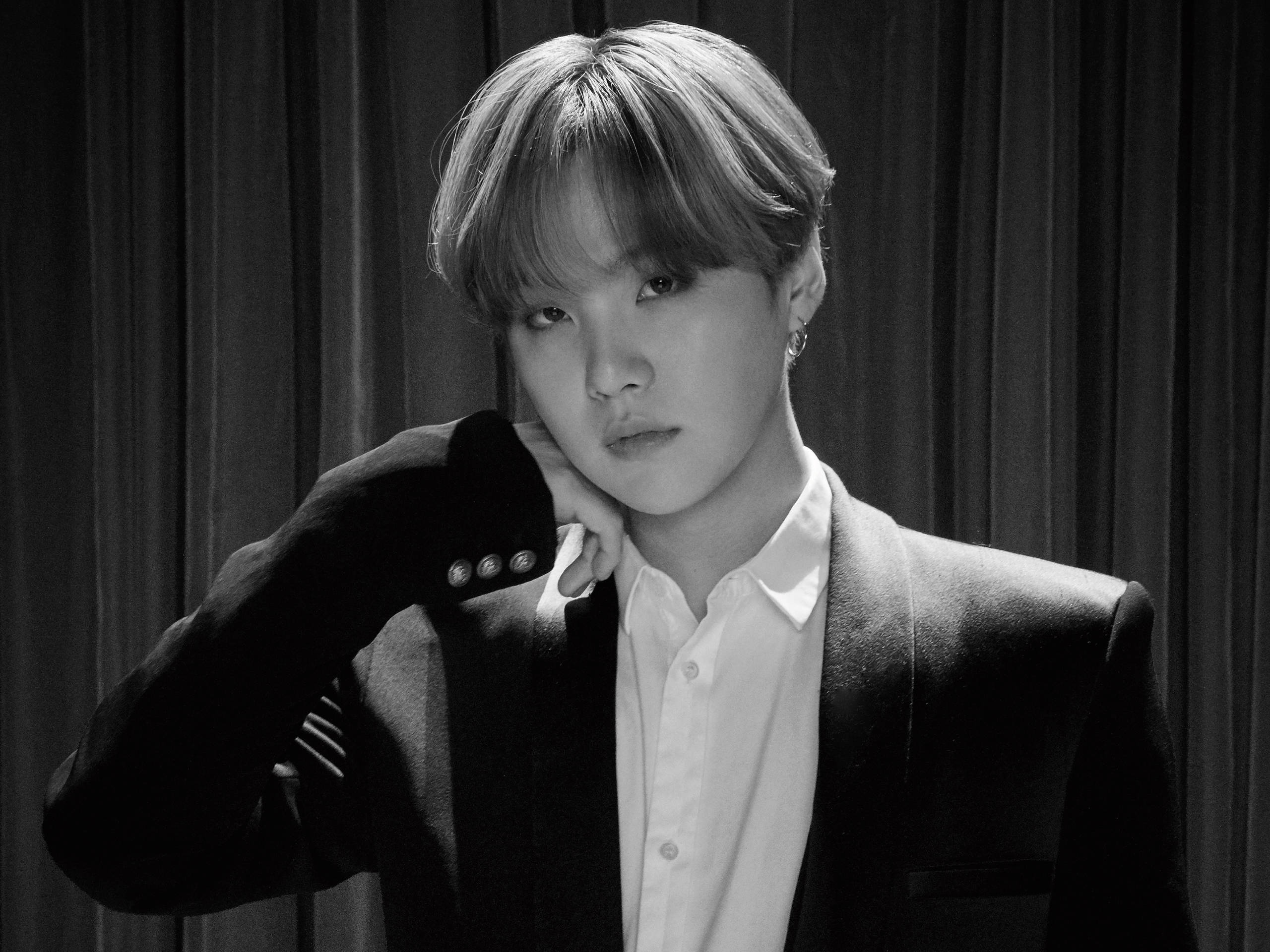 Suga BTS《Stay Gold》 壁紙 — 一個黑白肖像，穿著西裝的人將下巴擱在手上，背景是垂褶窗簾。