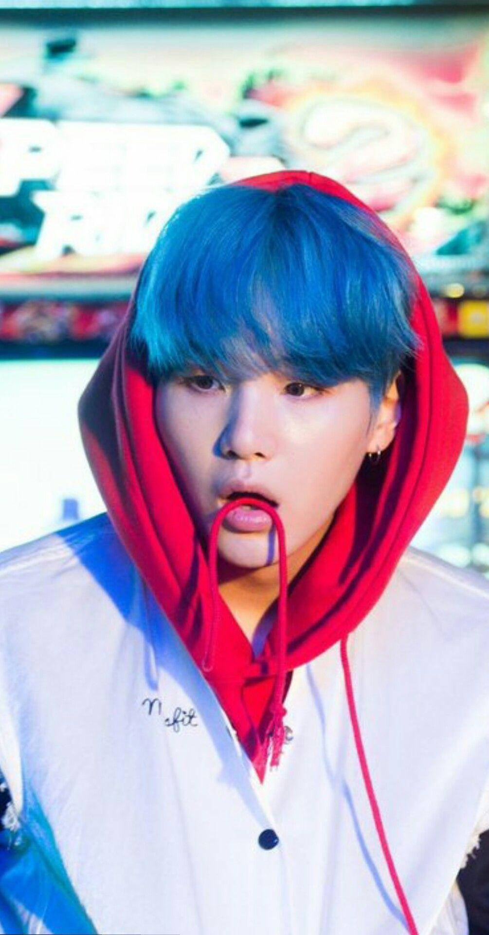 Suga BTS藍髮造型 壁紙 — 一個擁有鮮豔藍色頭髮的人，穿著紅色連帽衫和白色外套，背景是色彩繽紛且模糊的。