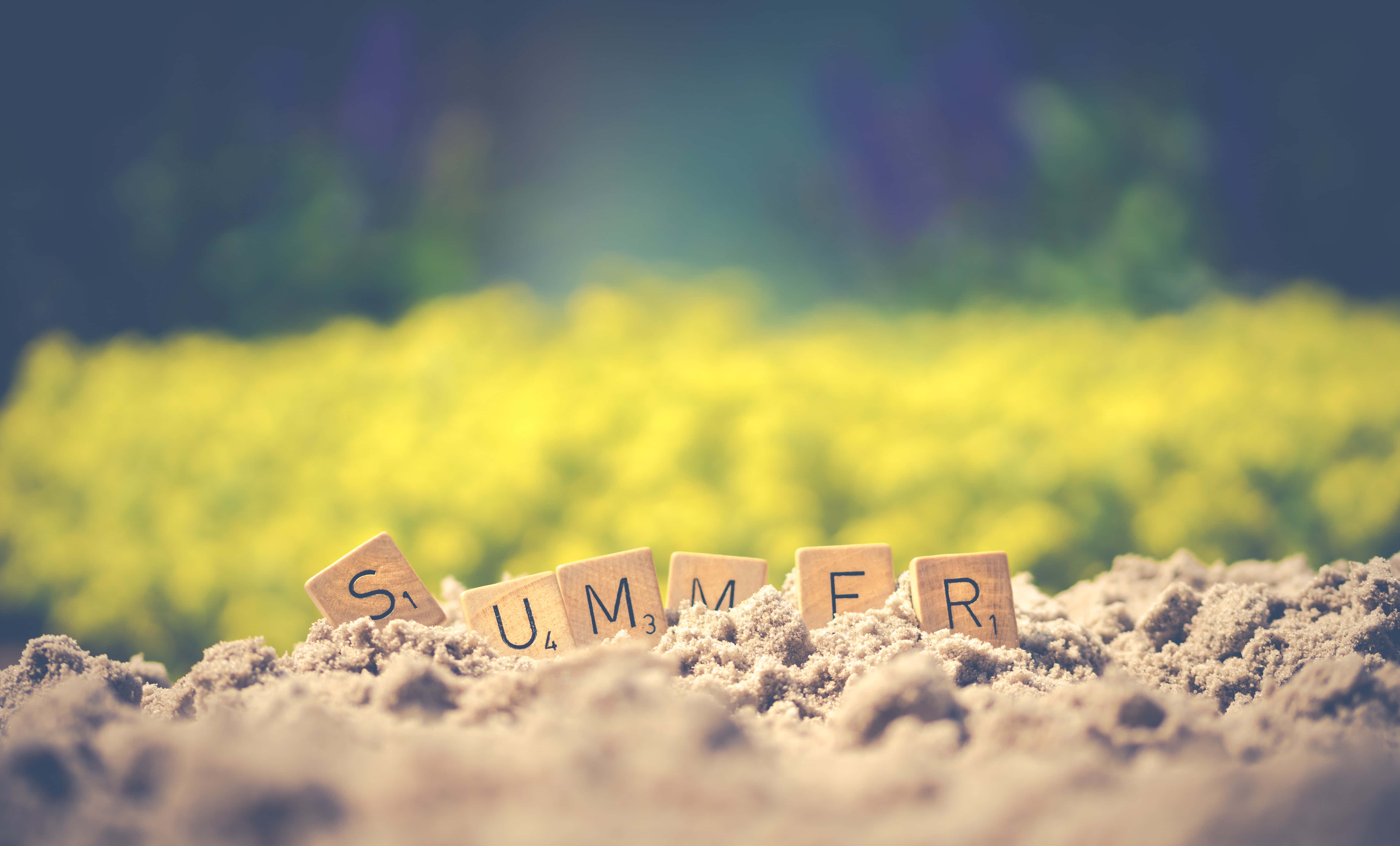 拼字遊戲方塊拼出「SUMMER」 壁紙 — 拼出夏天的木製字母瓷磚從沙質土壤中浮現，背景是柔和焦點的鮮黃色花田。