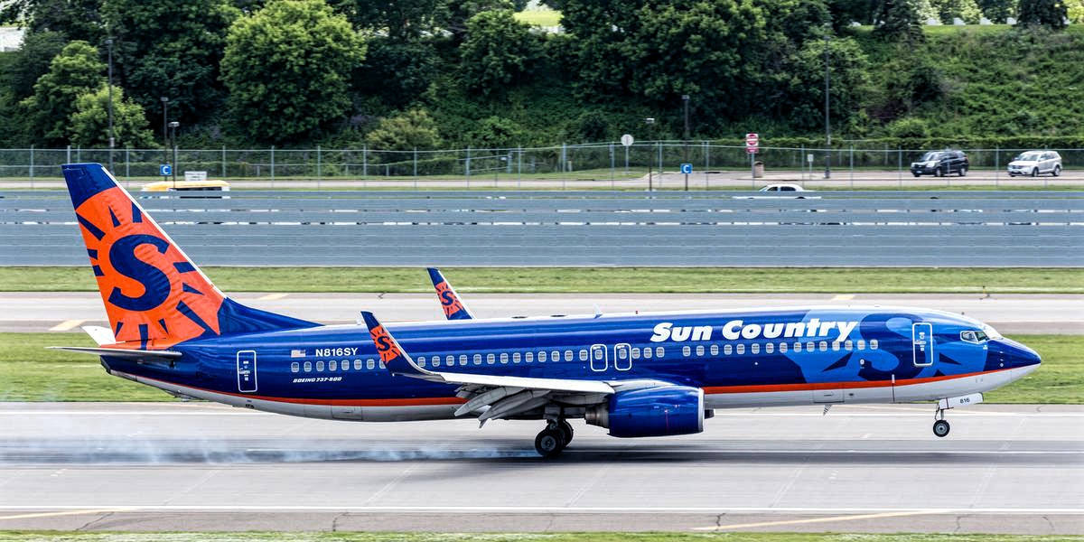 Sun Country航空波音737-8Q8客機起飛 壁紙 — 一架 Sun Country Airlines 的波音 737 在跑道上滑行，背景有茂密的綠樹和遠處的汽車。