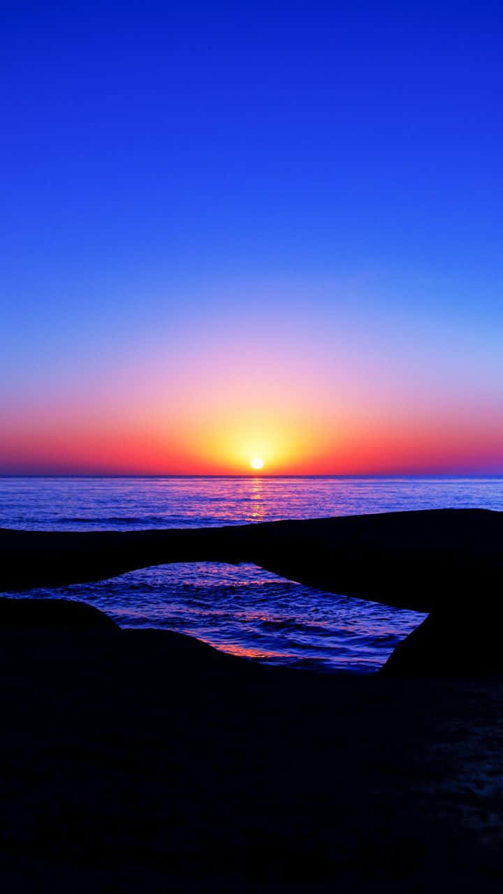 Coastal Sunset 桌布 壁紙 — 夕陽將天空染成火橙色和深藍色，太陽在平靜的海面上沉入地平線，四周被深色岩石剪影環繞。