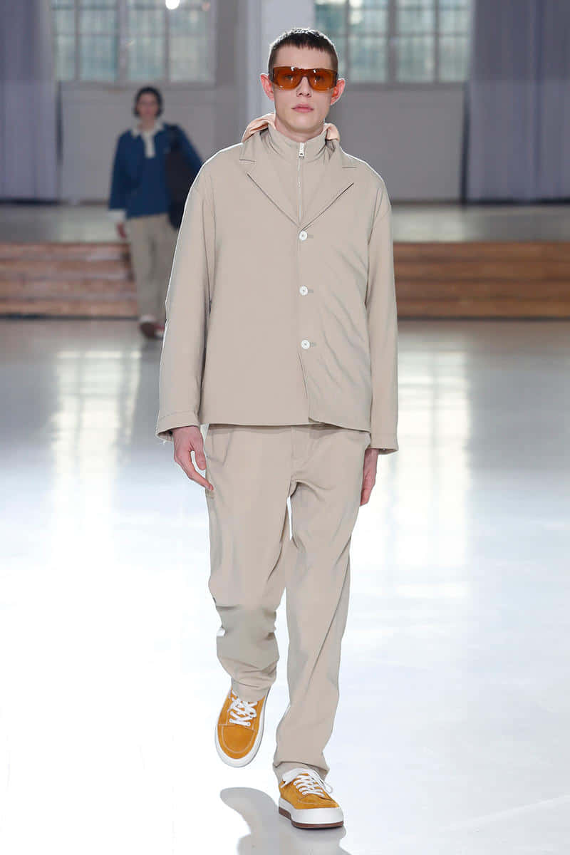Sunnei 2019 秋冬系列 Look 1 壁紙 — 一位男模特兒自信地走在明亮的時裝展台上，身穿米色西裝，戴著橙色太陽眼鏡，腳穿黃色運動鞋。