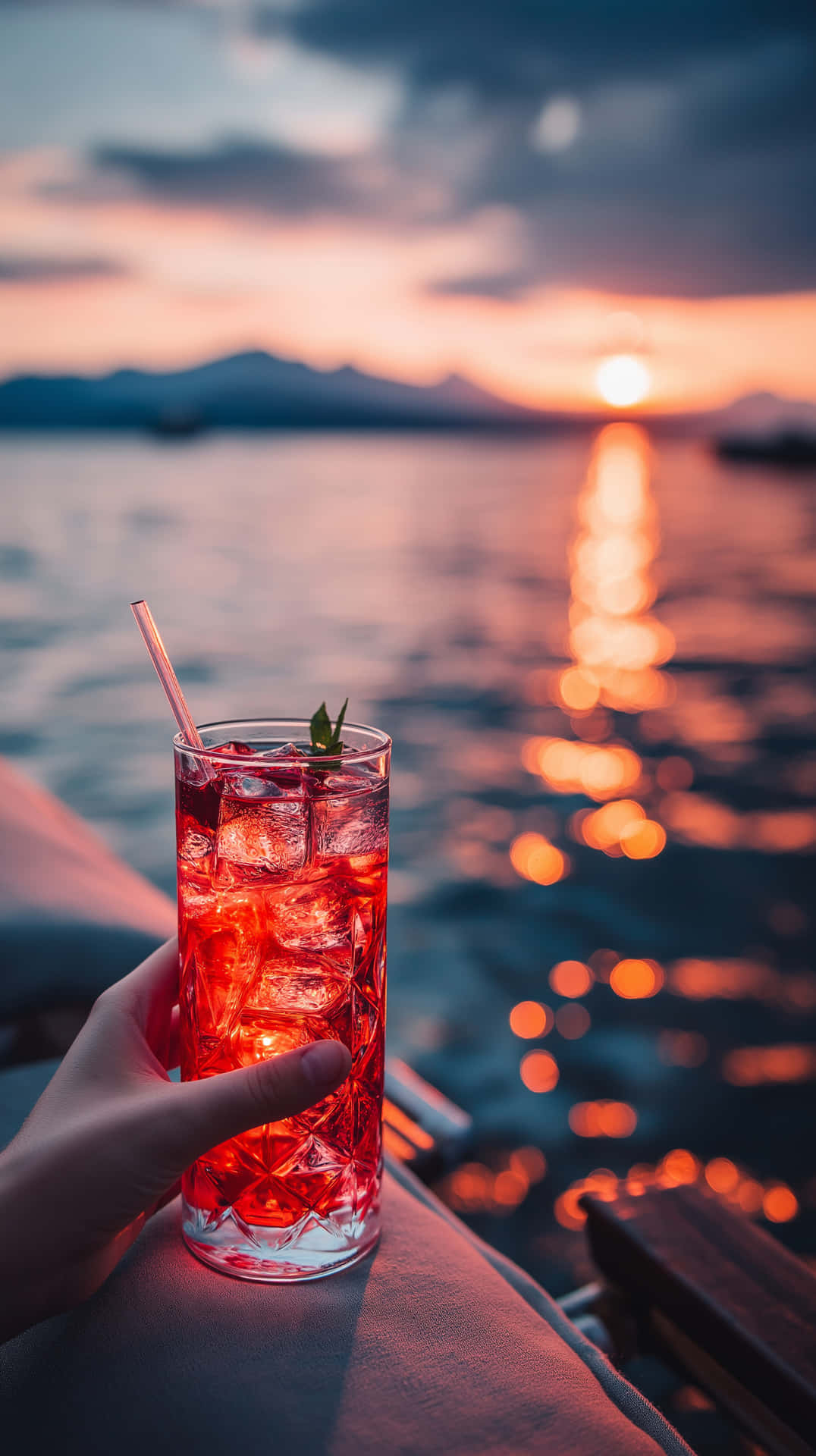 手拿紅色雞尾酒杯，加冰和薄荷，夕陽映照水面，靠近山巒