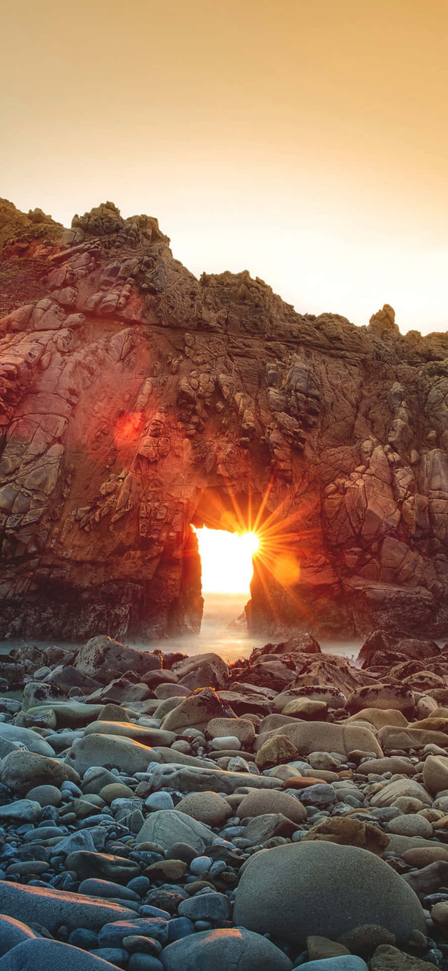  壁紙 — 夕陽光芒穿過天然岩石拱門，灑在卵石海灘上，呈現溫暖的金色色調。