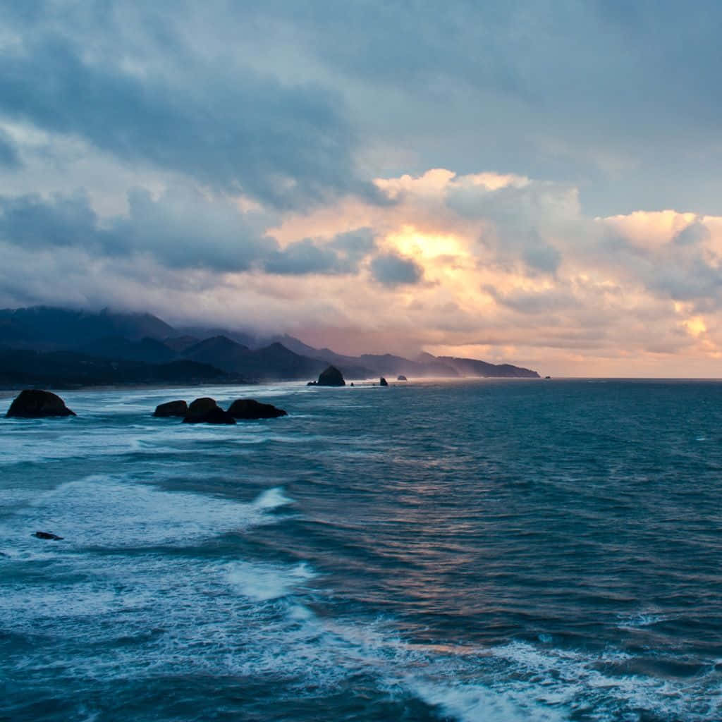 Coastal Sunset 桌布 壁紙 — 夕陽將雲彩染成金色，映照在波濤洶湧的海洋上，遠處有深色岩石和霧氣繚繞的山巒。