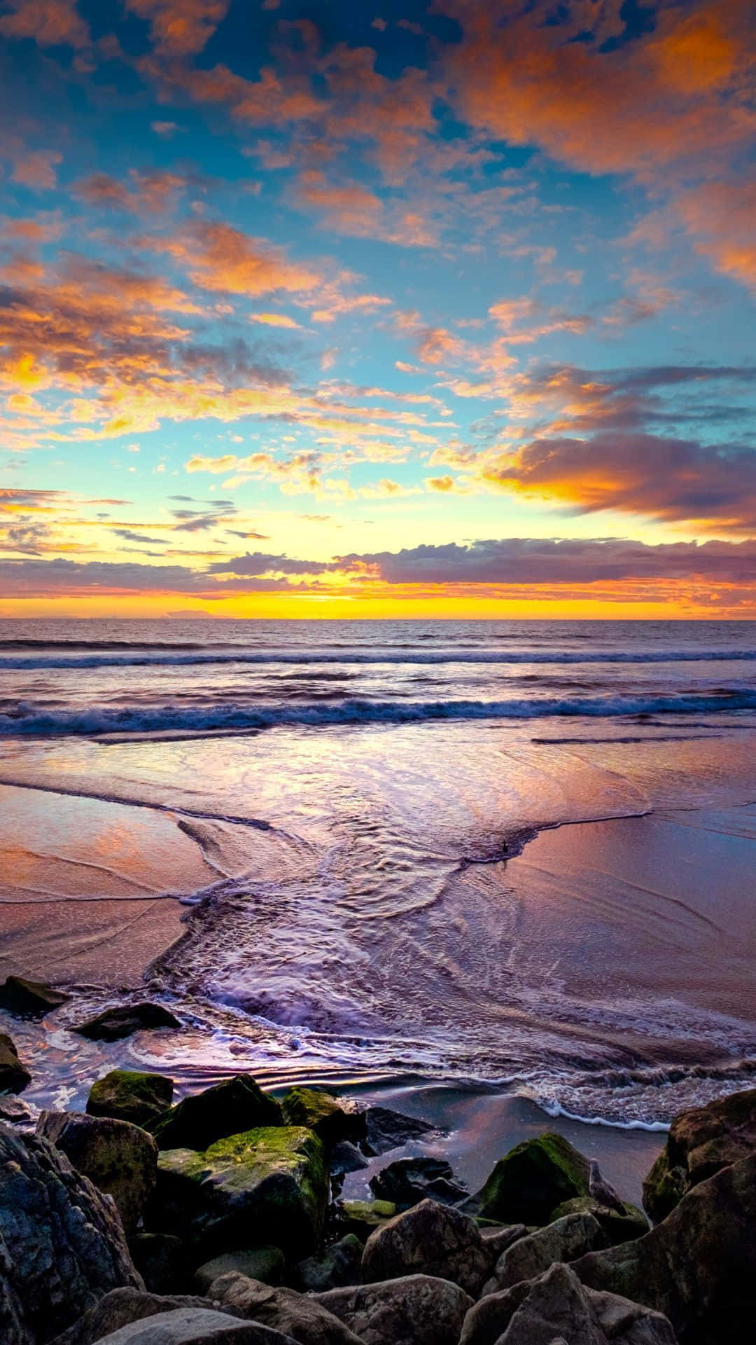 Coastal Sunset 桌布 壁紙 — 夕陽將天空染成火熱的橙色和藍色，海浪輕輕拍打著佈滿苔蘚岩石的沙灘。